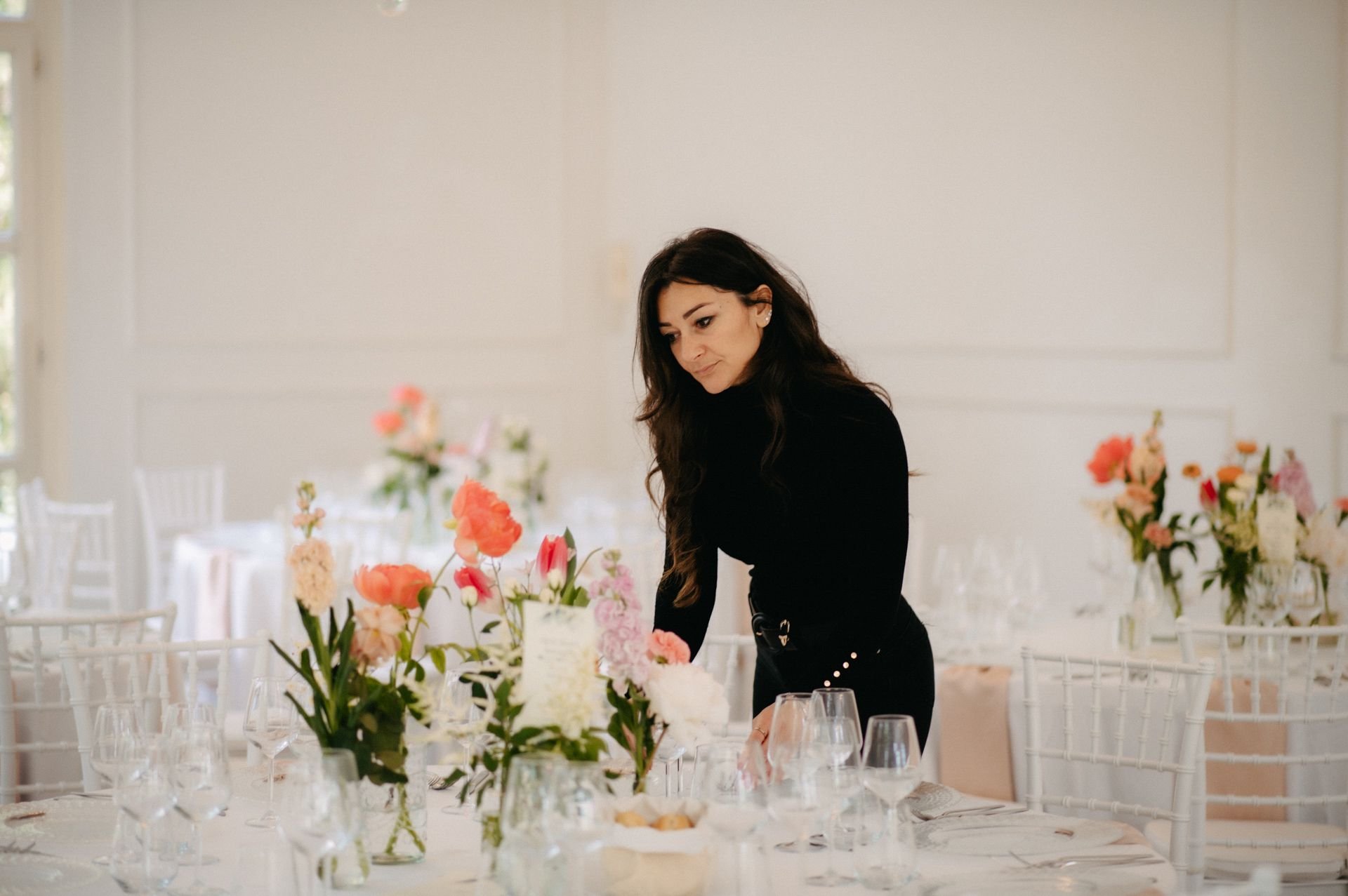 A woman arranging flowers on a table in a bright, elegant room decorated with floral centerpieces and set for a formal event.