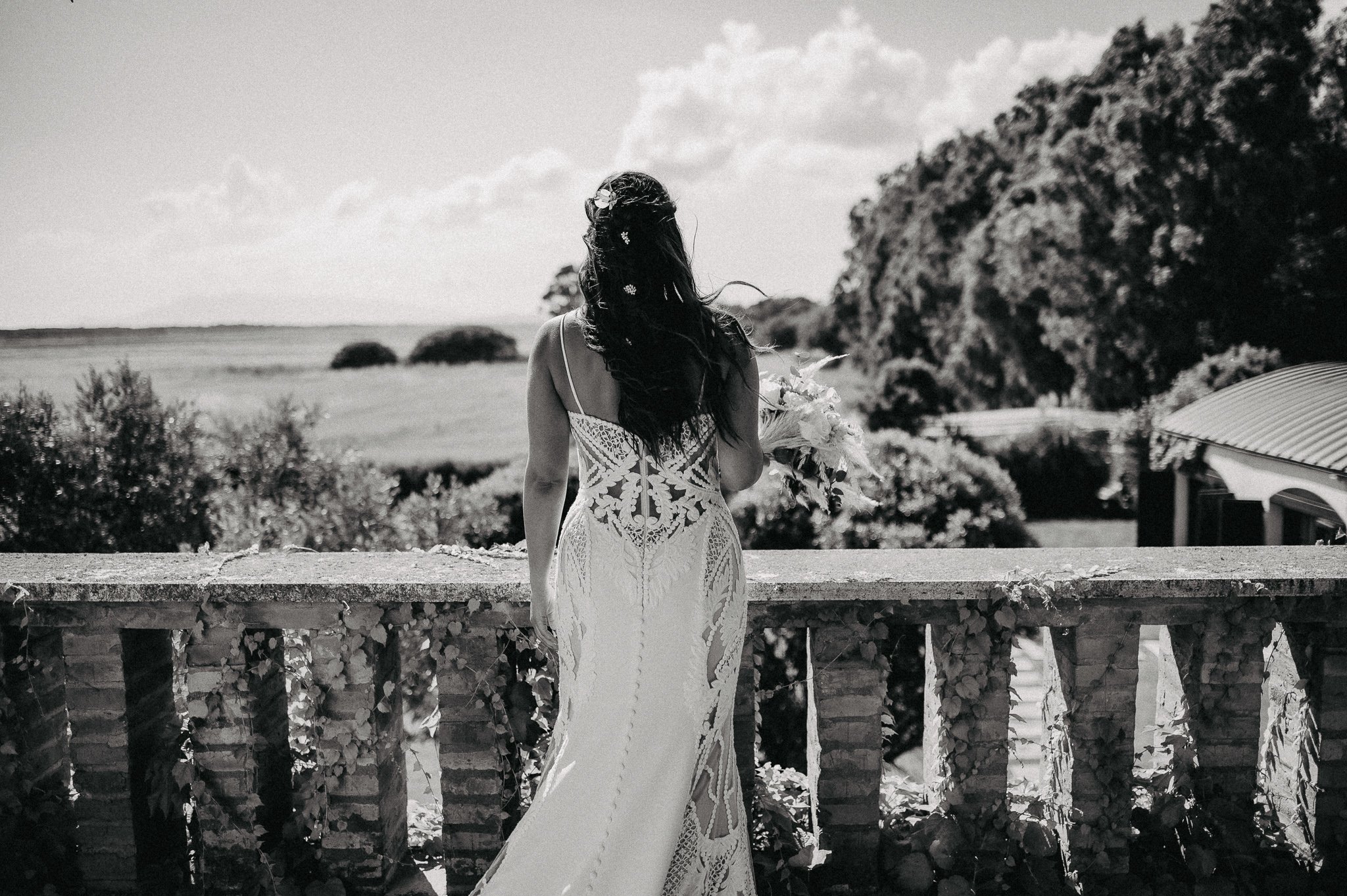 A woman in a white lace dress holding a bouquet of flowers, standing on a balcony overlooking a rural landscape with trees and open sky.