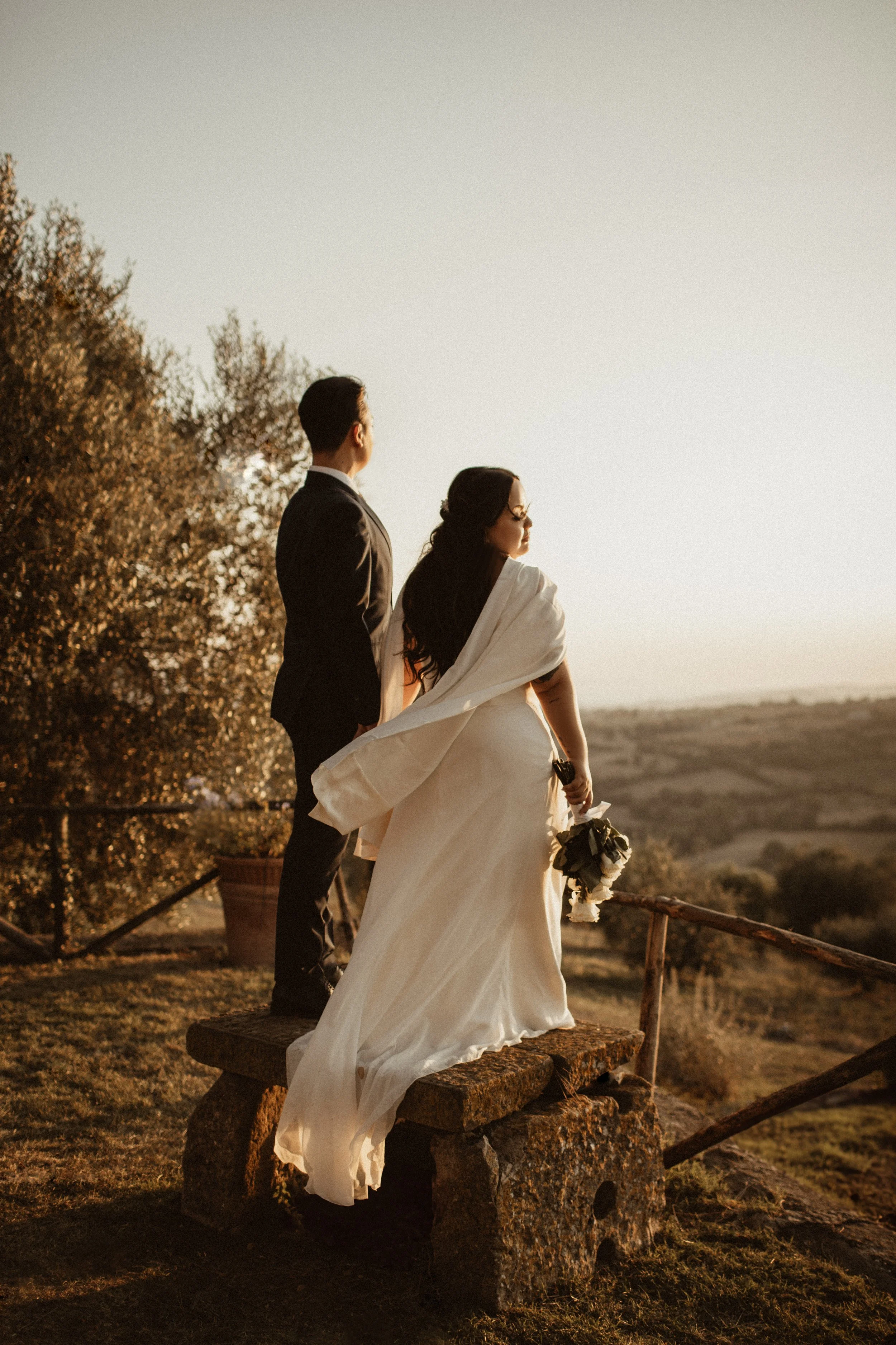 A bride and groom standing on a stone bench outdoors during sunset, with the bride holding a bouquet and the groom looking into the distance.