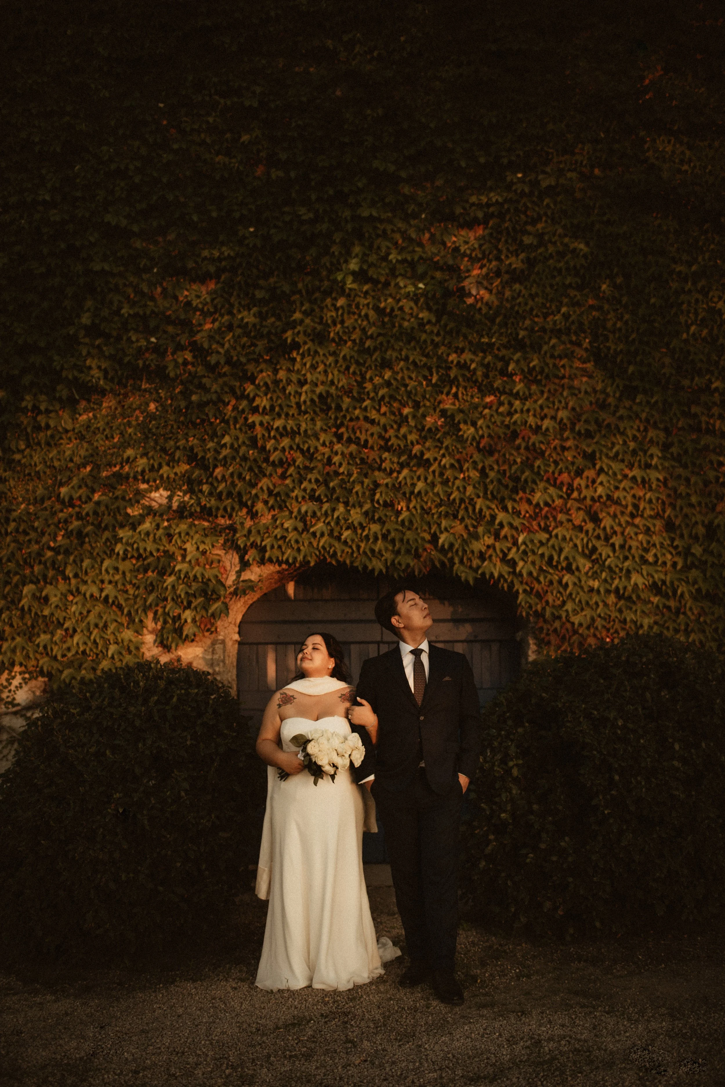 Photo of a bride and groom standing outdoors at night, holding hands, with a large tree with green leaves in the background.