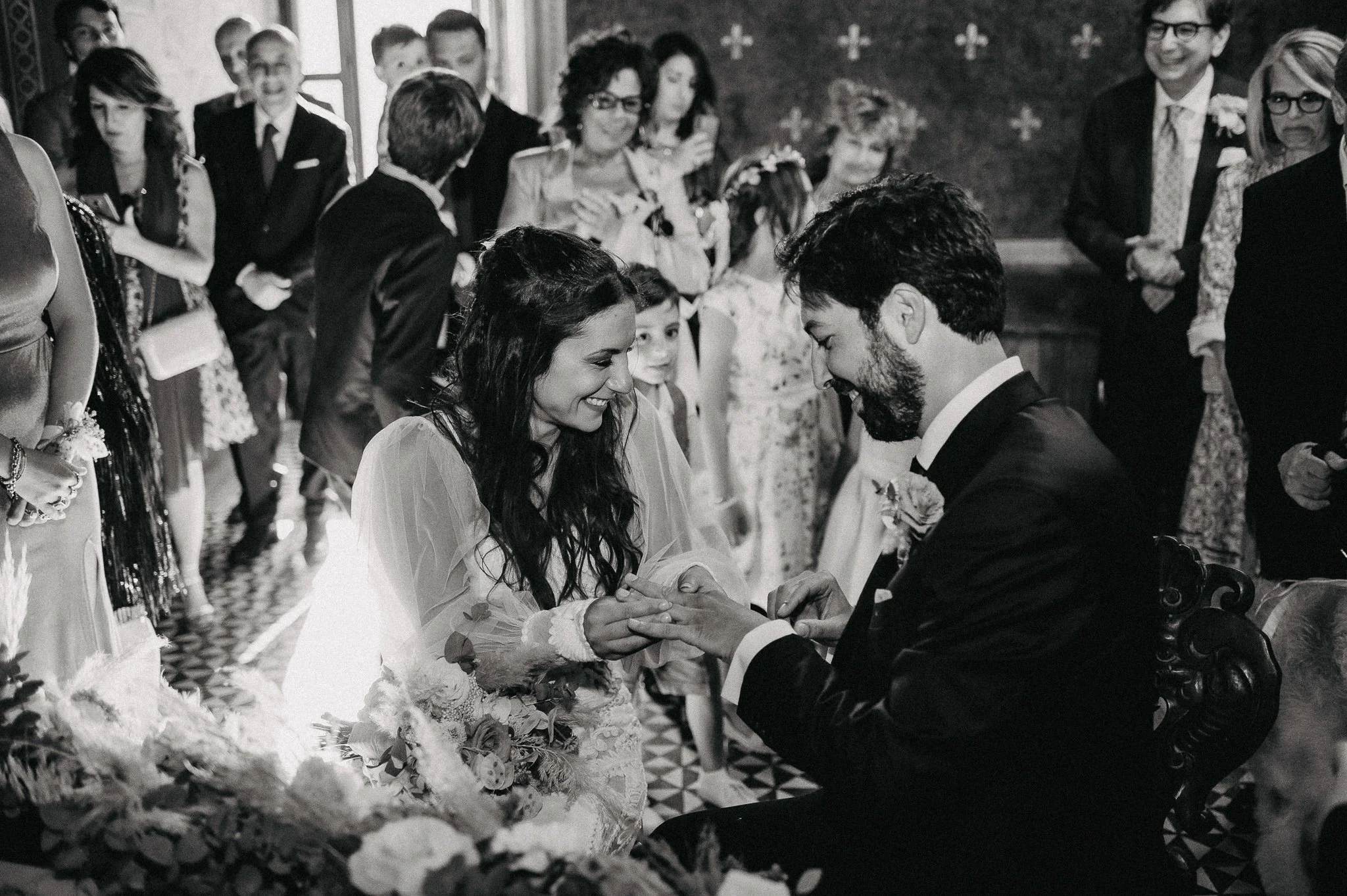 A wedding ceremony with a bride and groom exchanging rings, surrounded by family and friends in a decorated indoor setting.