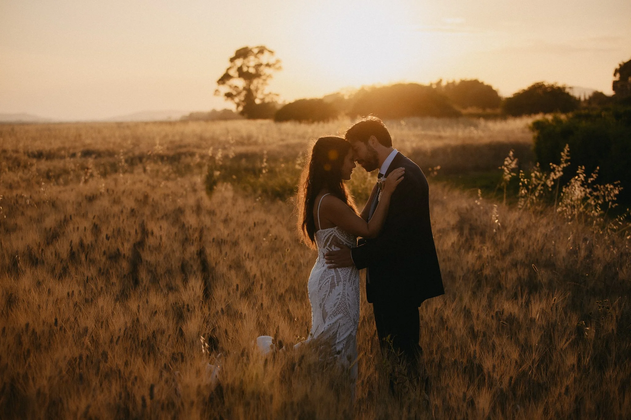 A newlywed couple embracing in a field at sunset, with a woman in a white lace dress and a man in a black suit, sharing an intimate moment as the sun sets behind them.