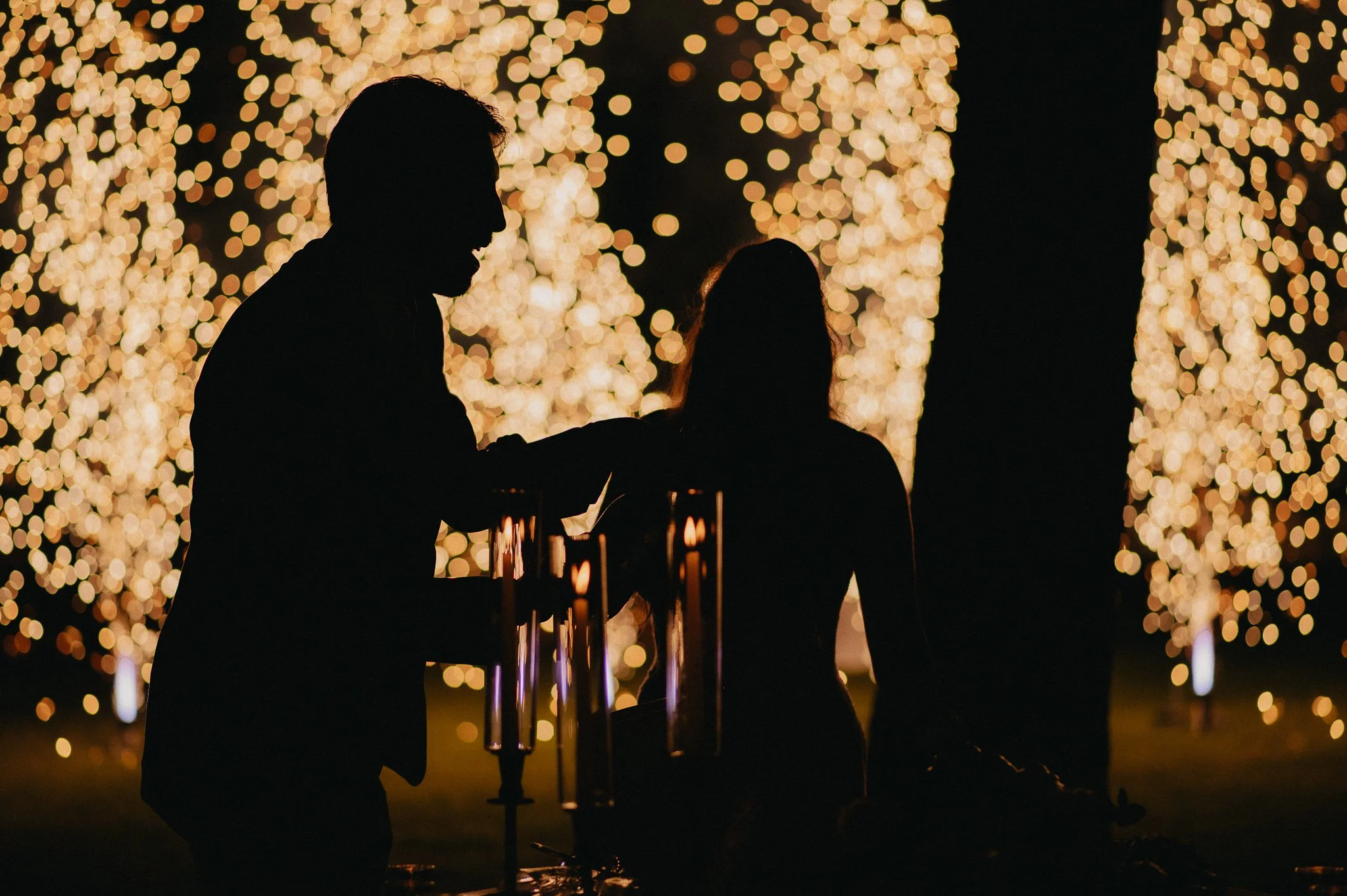 Silhouettes of two people at a nighttime outdoor event with blurred string lights in warm tones in the background.