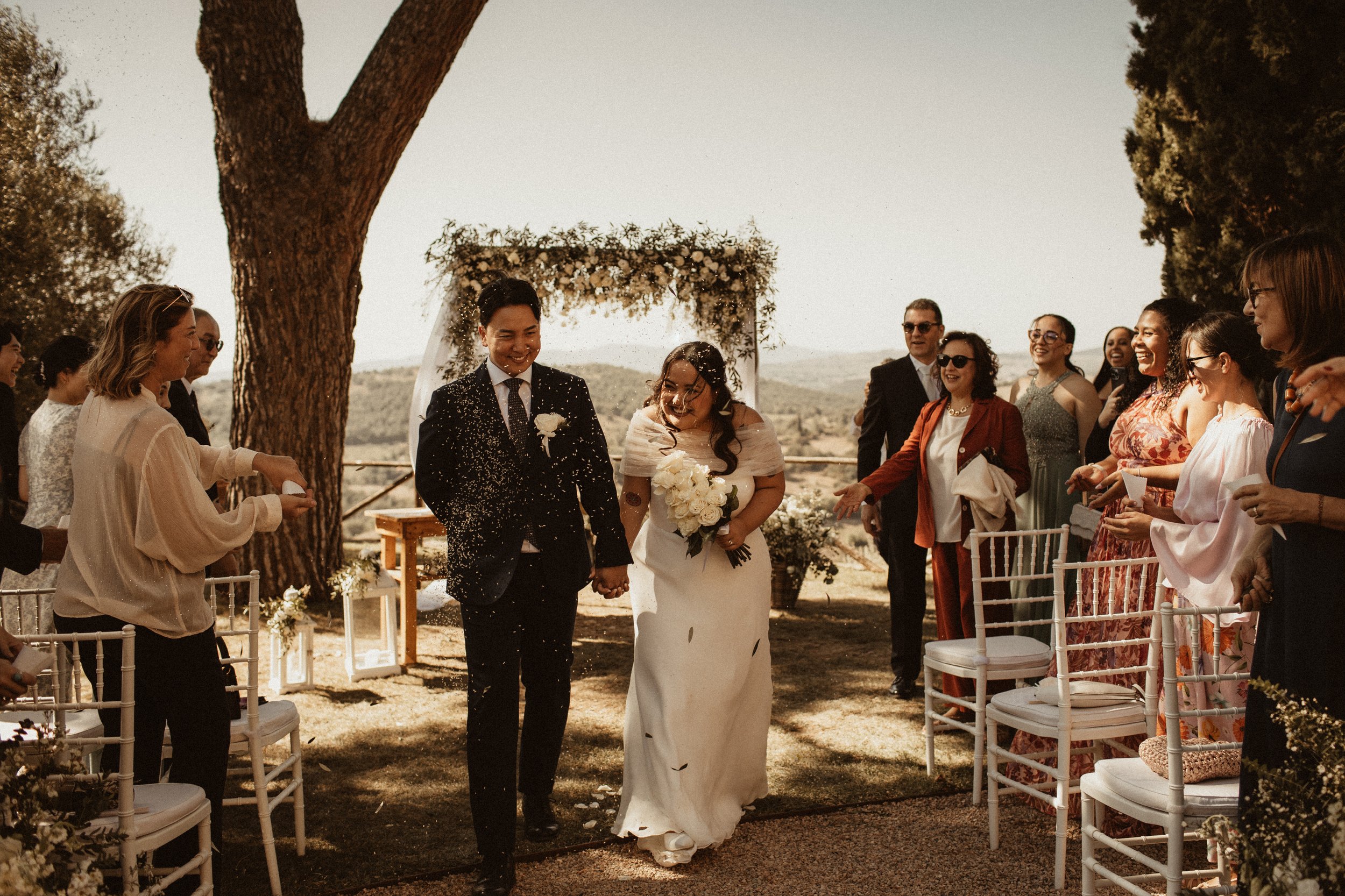 A bride and groom walk hand in hand down an outdoor aisle, smiling, as guests celebrate at a wedding reception.
