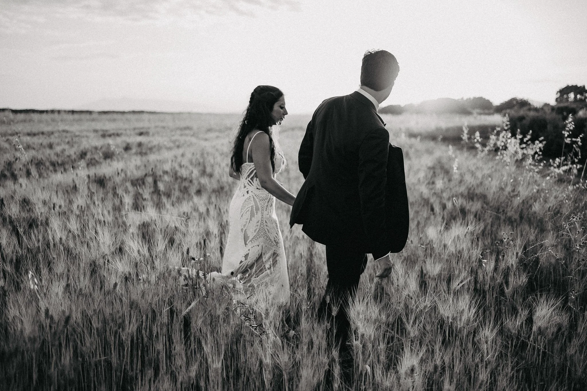 A black-and-white photo of a woman in a wedding dress and a man in a suit walking through a field during sunset.