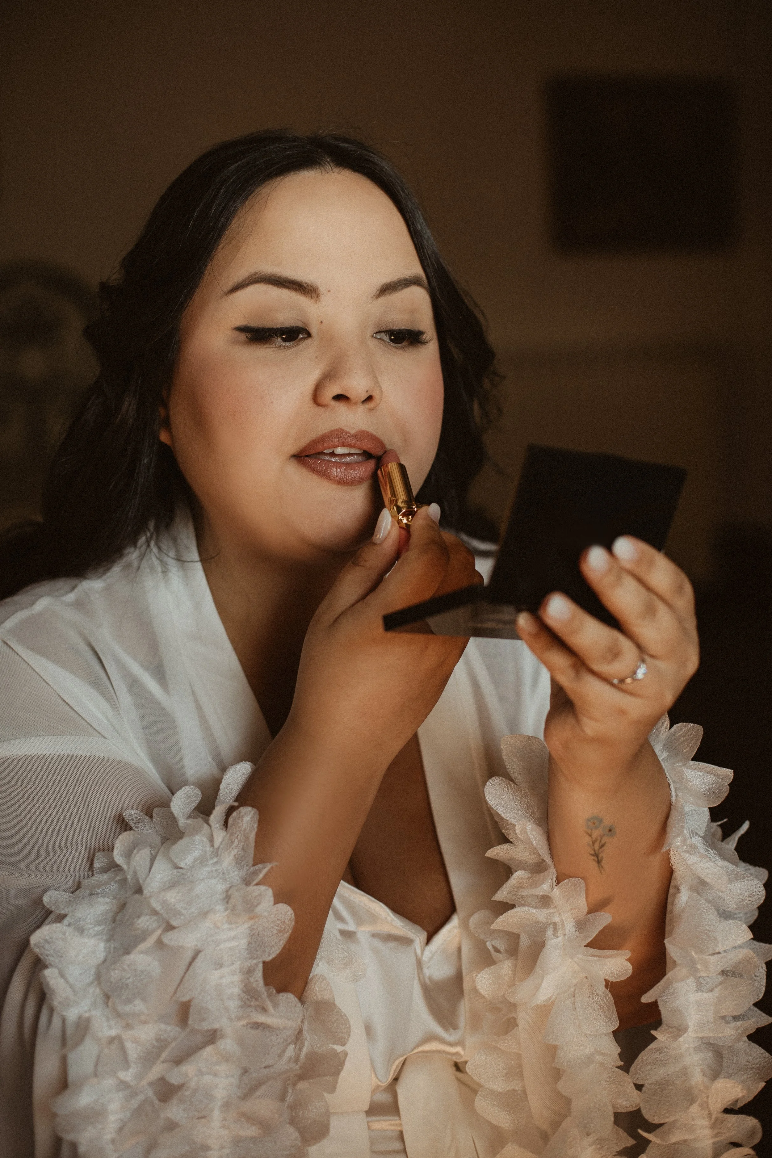 Woman applying lipstick while looking into a mirror, wearing a white garment with floral accents on the sleeves.