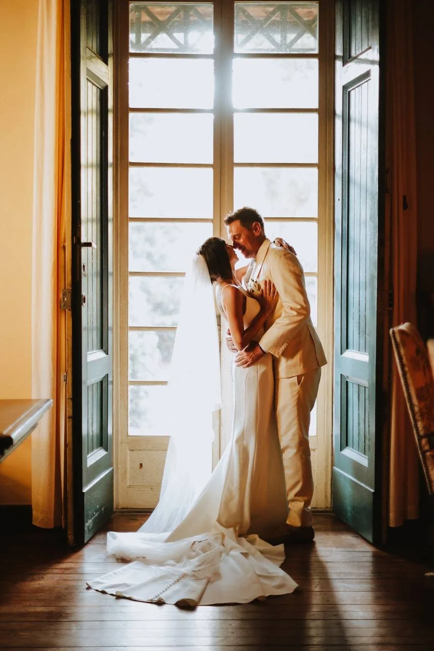 A bride and groom sharing a kiss in front of a large window with open green shutters, brightly lit from behind, inside a rustic room with wooden floors.