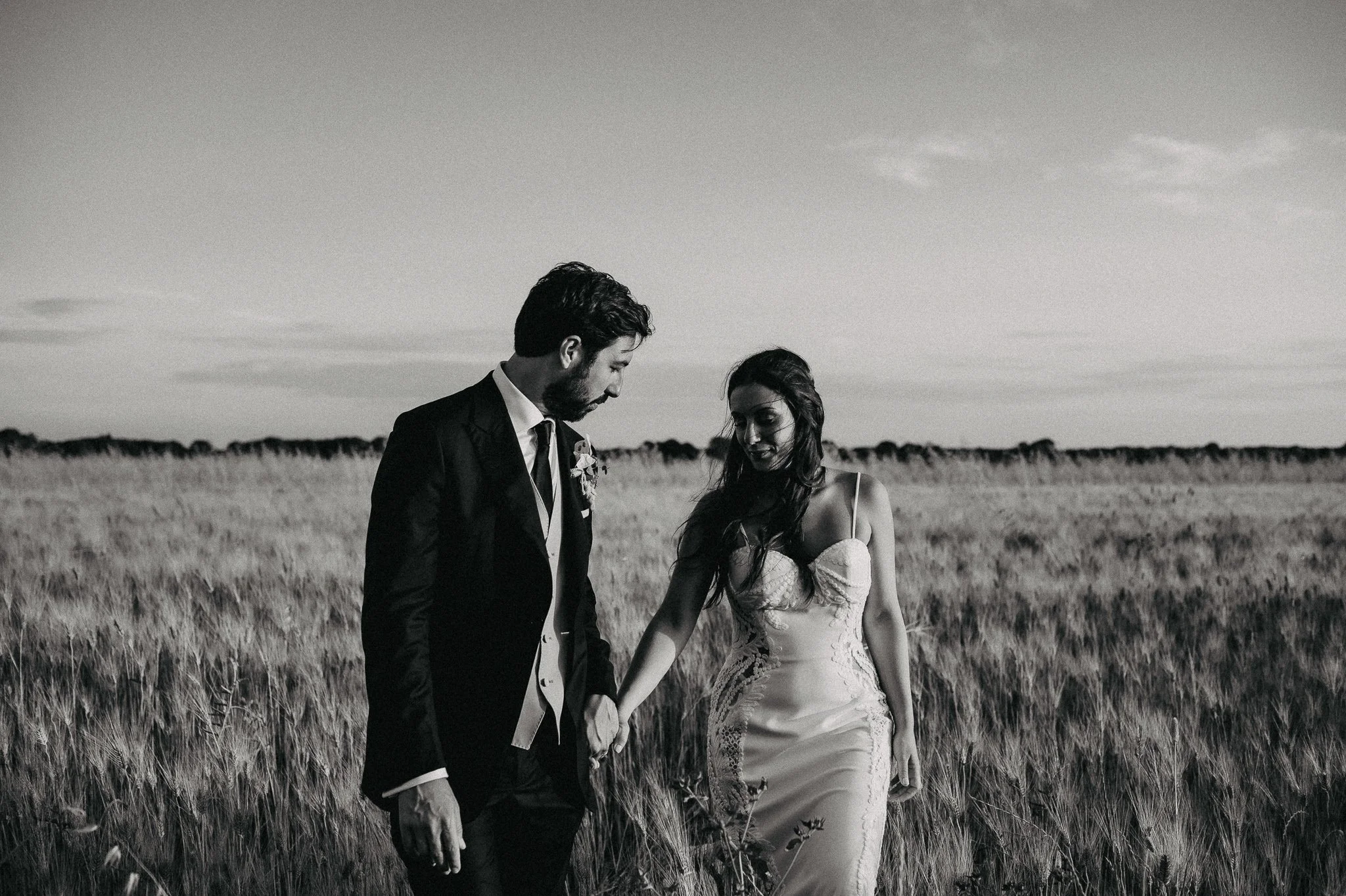 A black-and-white photo of a bride and groom holding hands in a field of tall grass, looking at each other, with a cloudy sky in the background.