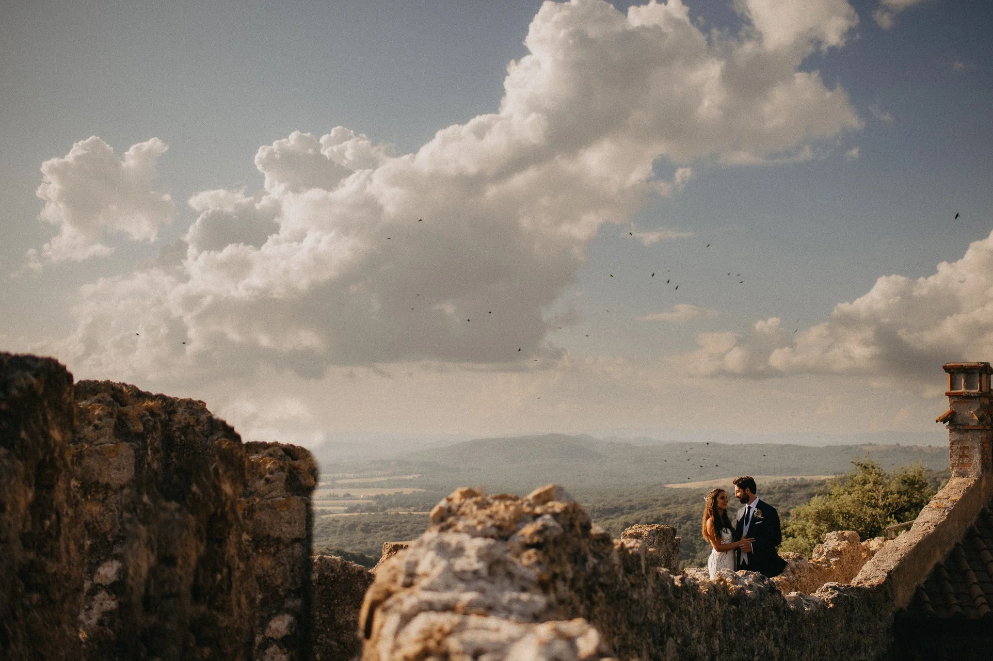 A couple dressed in wedding attire standing on a stone wall with a scenic landscape and a partly cloudy sky in the background.