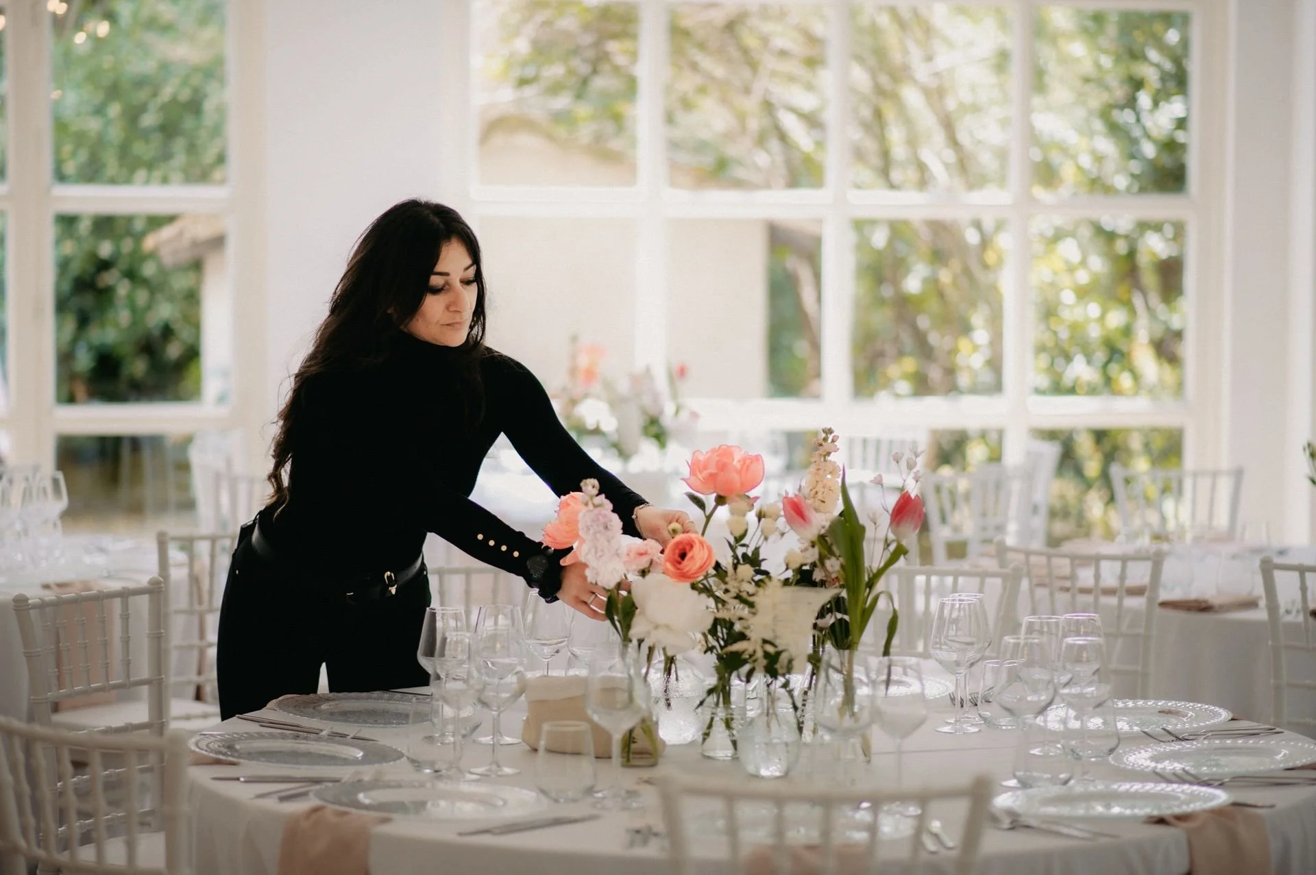 A woman arranging a floral centerpiece at a dining table in a bright, sunlit room with large windows.