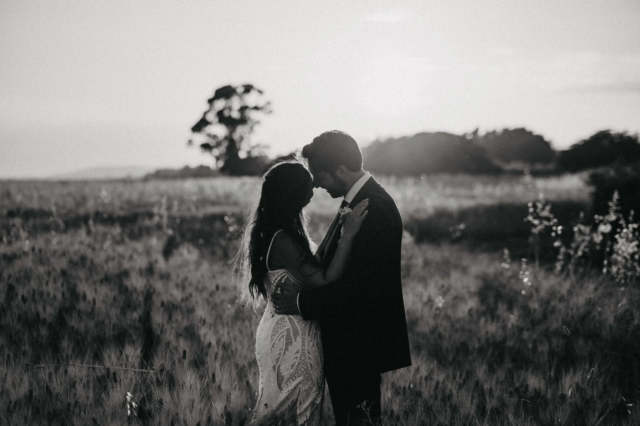 A black and white photo of a couple in wedding attire standing close together in a field at sunset, touching foreheads and embracing.