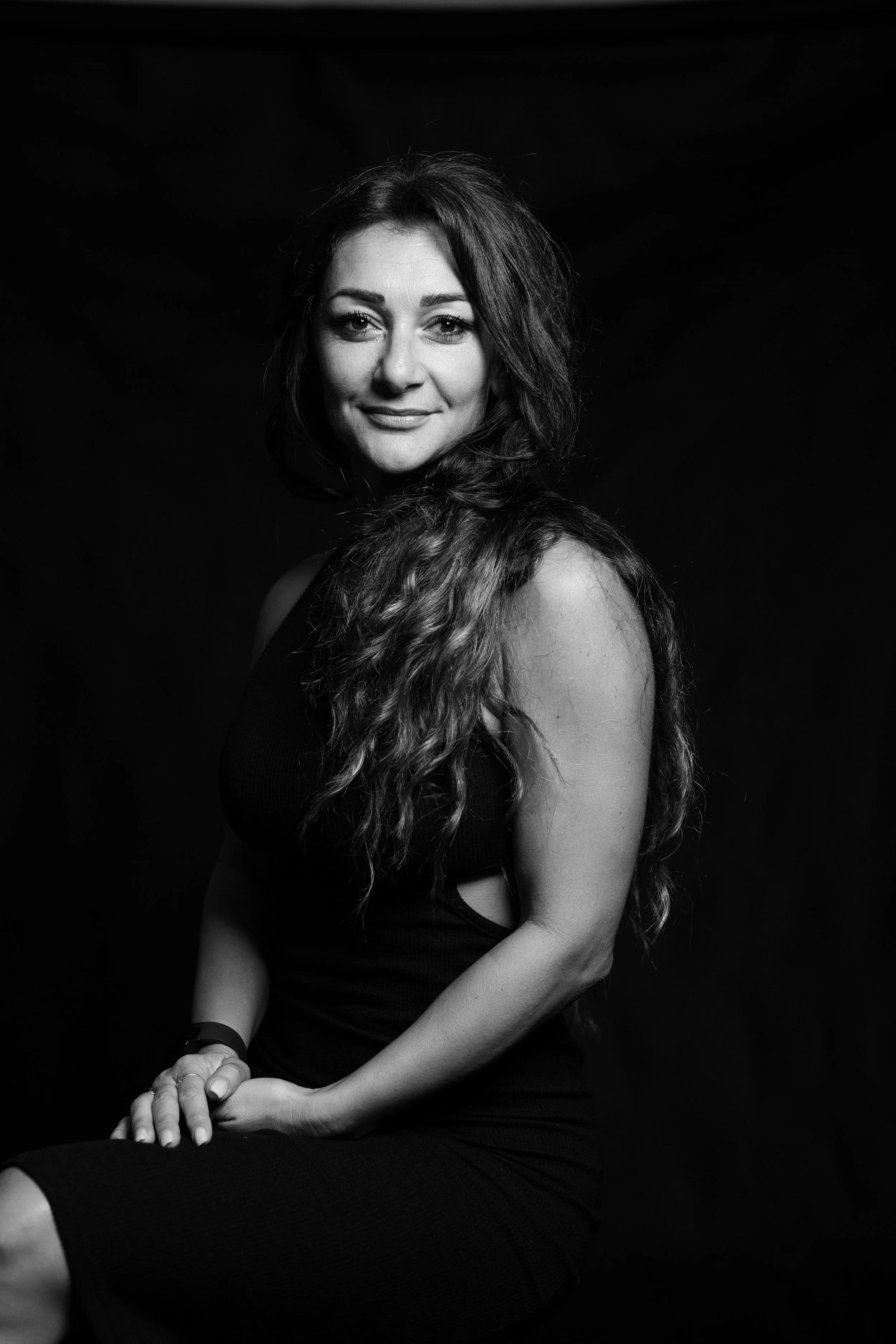 Black and white portrait of a woman with long, wavy hair, wearing a sleeveless dress, sitting against a dark background, smiling softly at the camera.