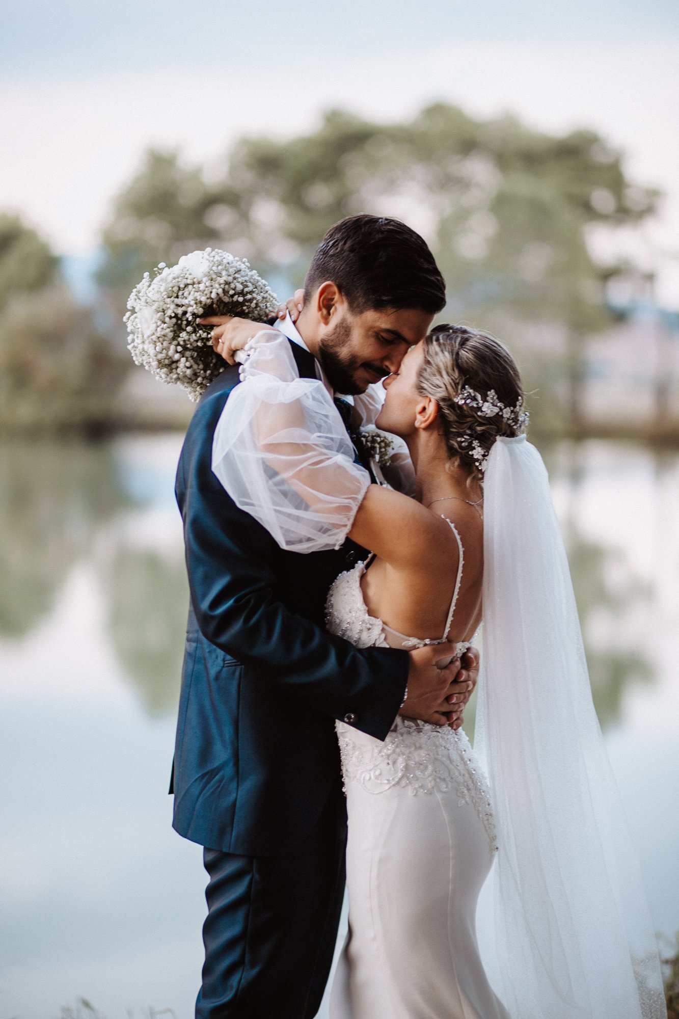 A bride and groom embracing outdoors near a lake during their wedding, with trees in the background.