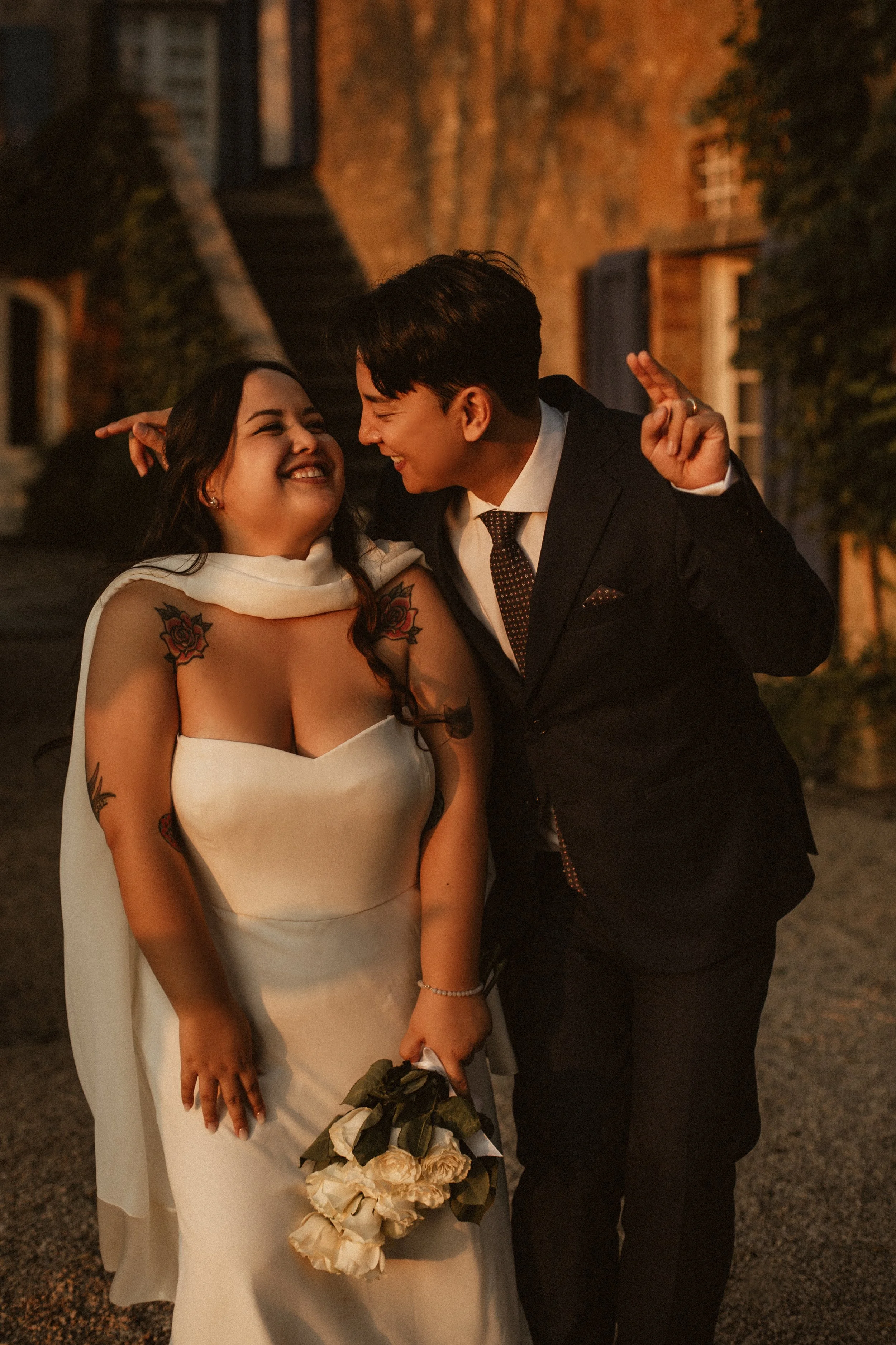 A bride and groom smiling and celebrating outdoors in the evening. The bride wears a white dress with tattoos on her arms and holds a bouquet of white roses. The groom is in a dark suit with a tie, leaning towards the bride with a joyful expression.