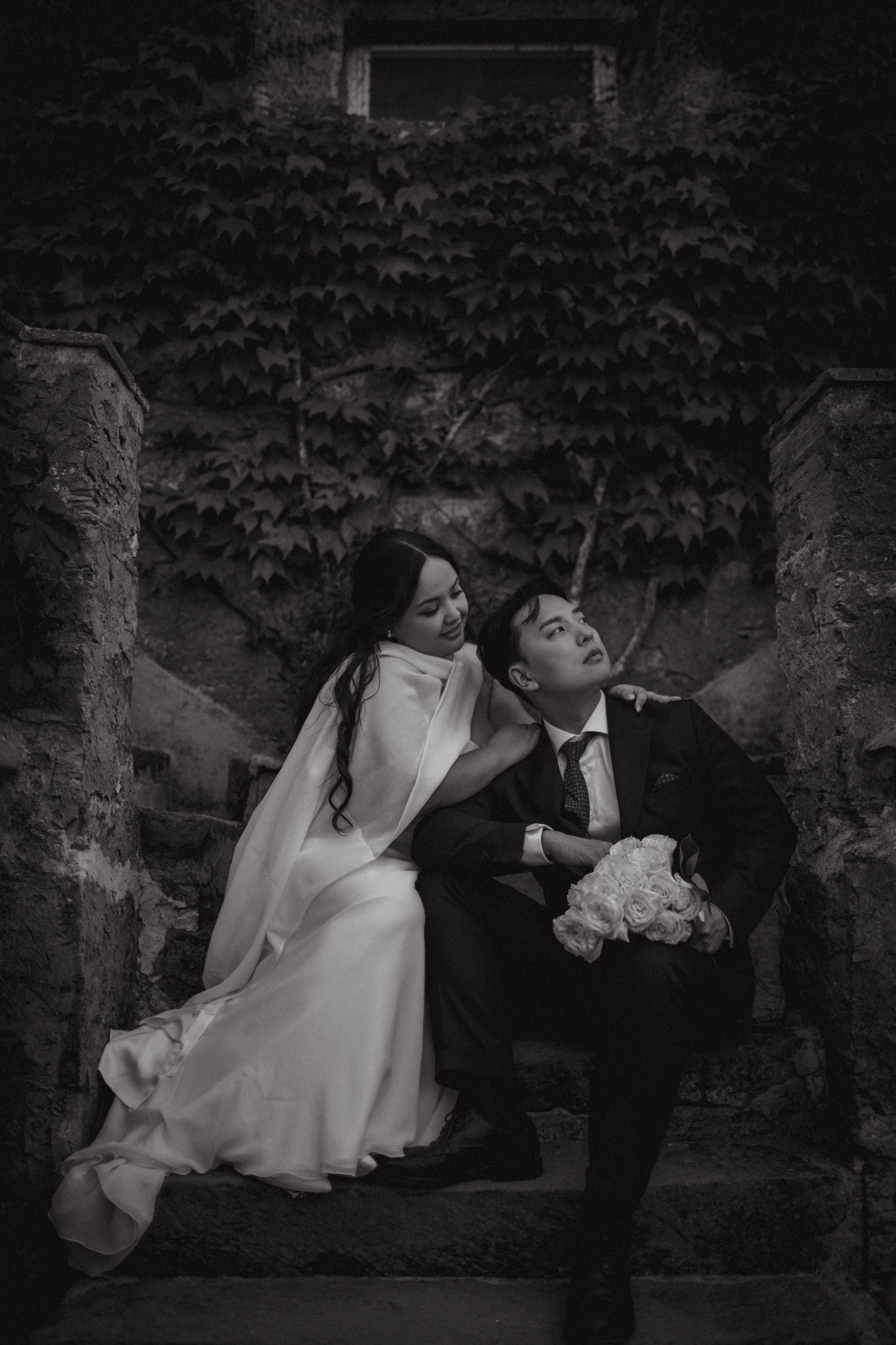 A black and white photo of a woman in a wedding dress and a man in a suit sitting on stone steps outside, with the woman leaning on the man and holding a bouquet of flowers, surrounded by a stone wall and ivy.