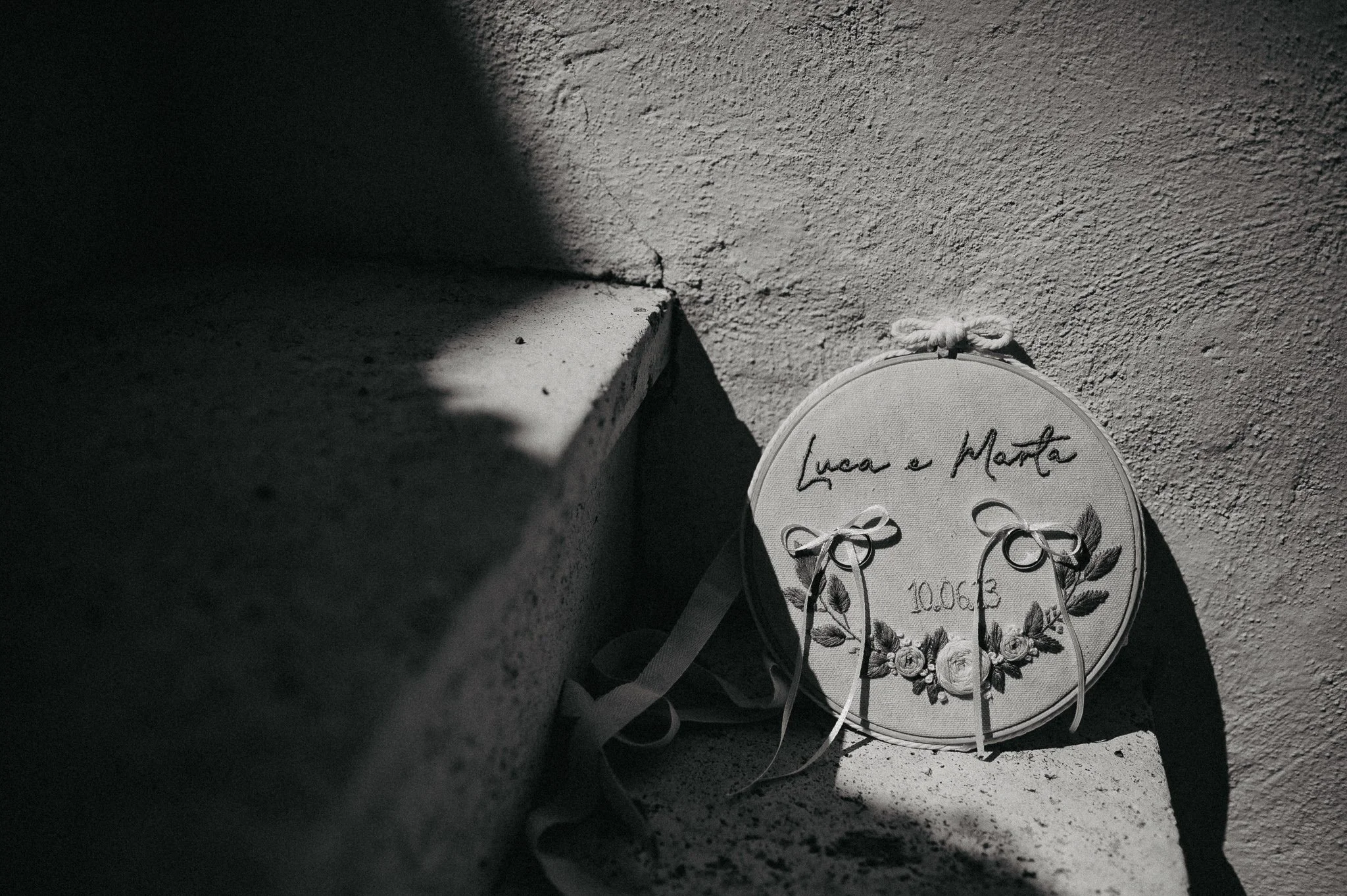 A round white embroidered bag with floral design and handwritten text leaning against a textured wall near a step, with sunlight casting shadows.