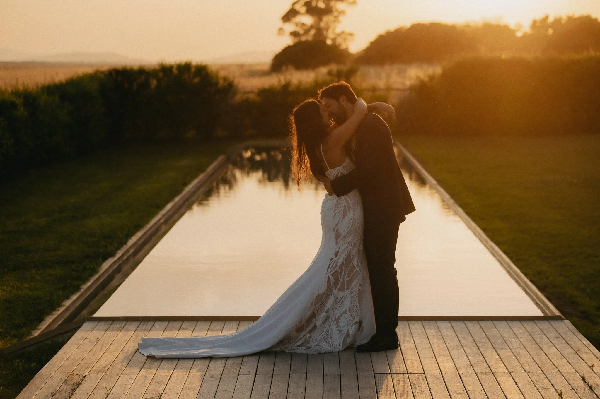 A couple in wedding attire sharing an intimate moment by a pool during sunset.