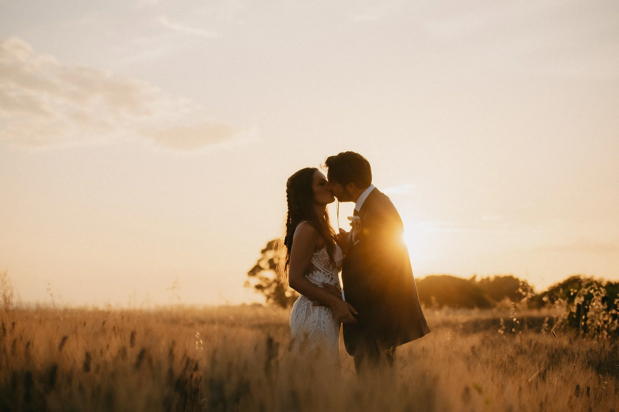 A couple kissing in a field during sunset, with the sun low in the sky creating a warm glow around them.