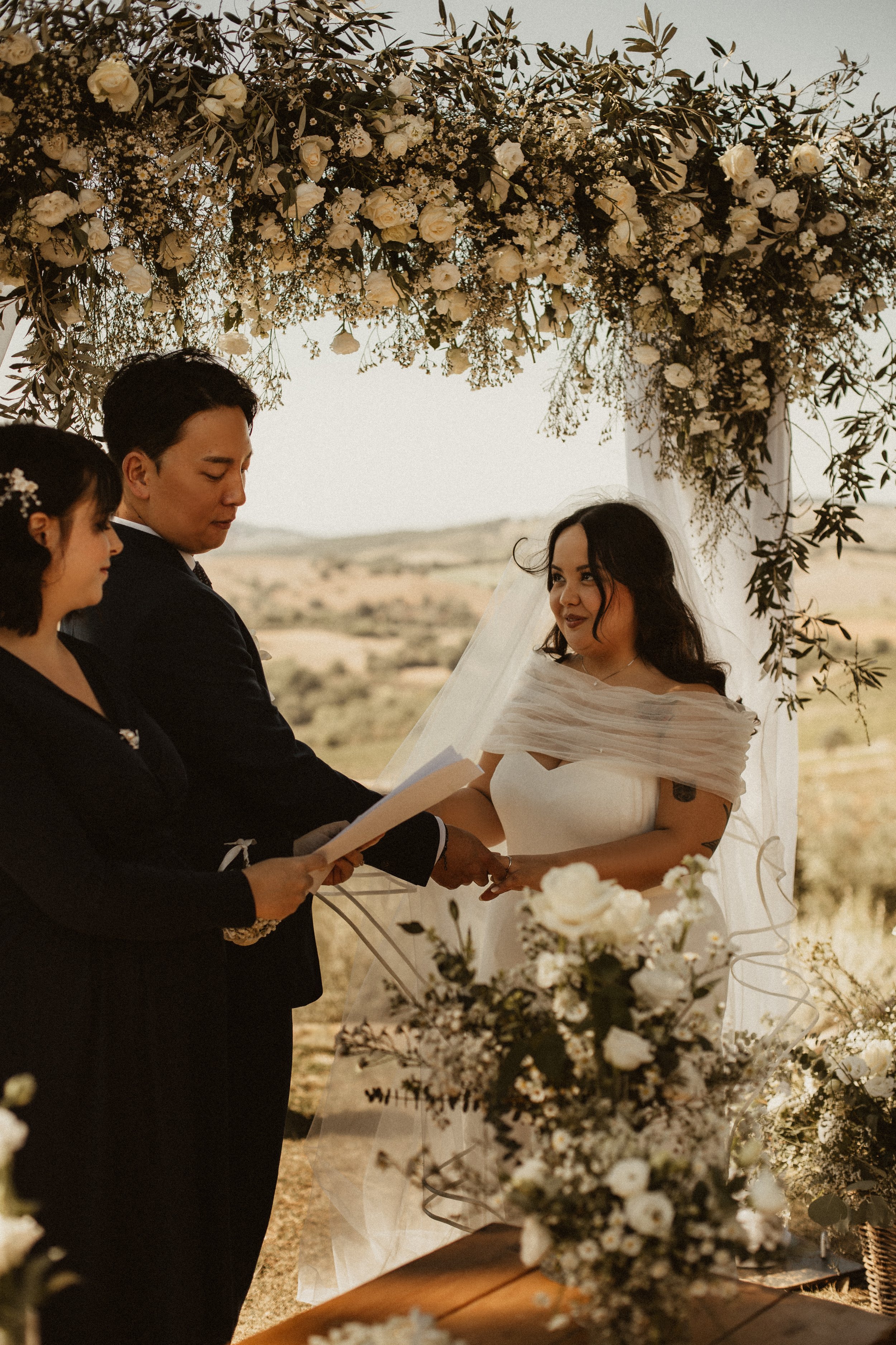 A wedding ceremony taking place outdoors with a bride and groom holding hands, standing under a floral arch with white roses and greenery, as an officiant reads from a paper.