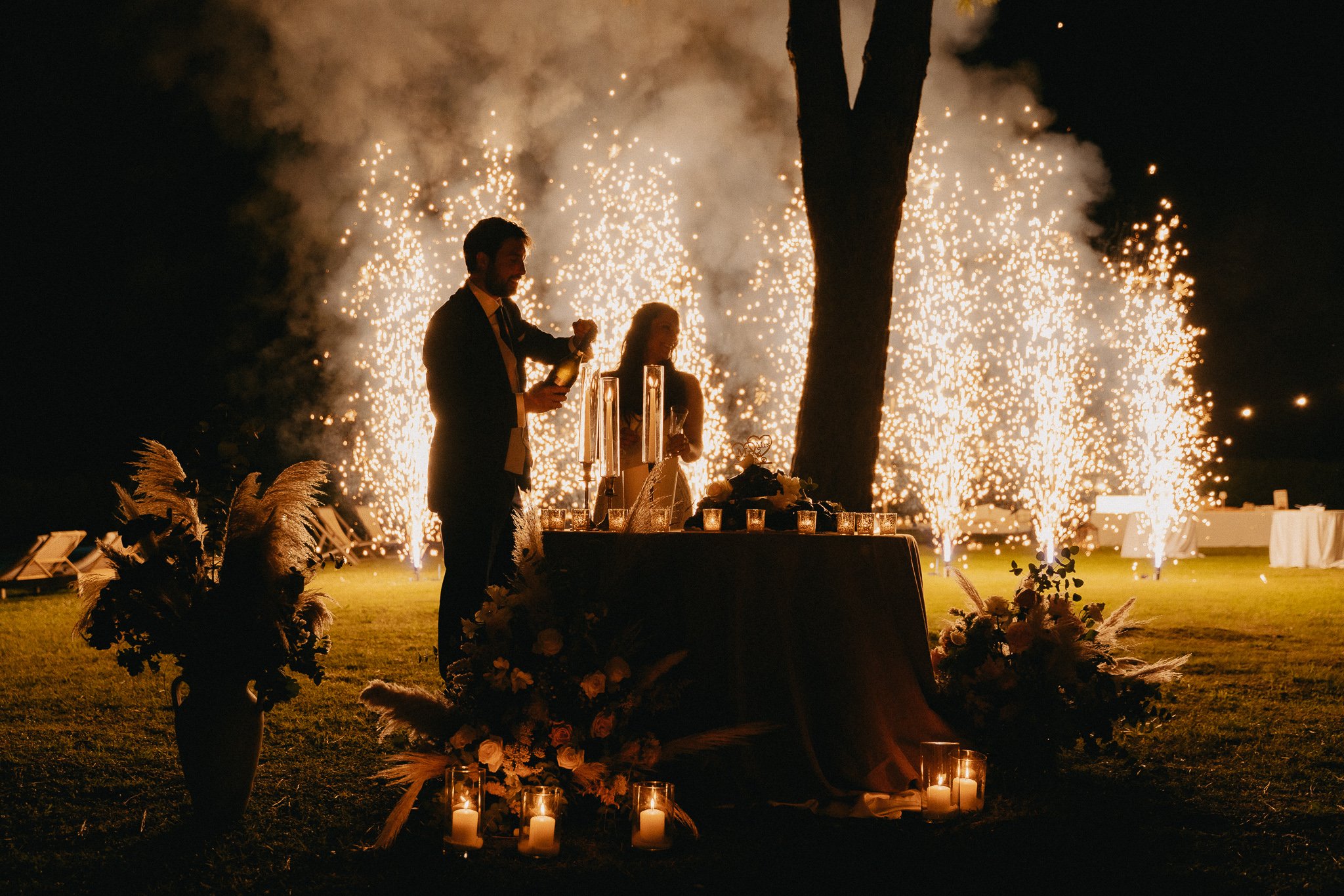 A couple at a wedding celebration cutting a cake under fireworks display at night, with a table decorated with candles and floral arrangements.