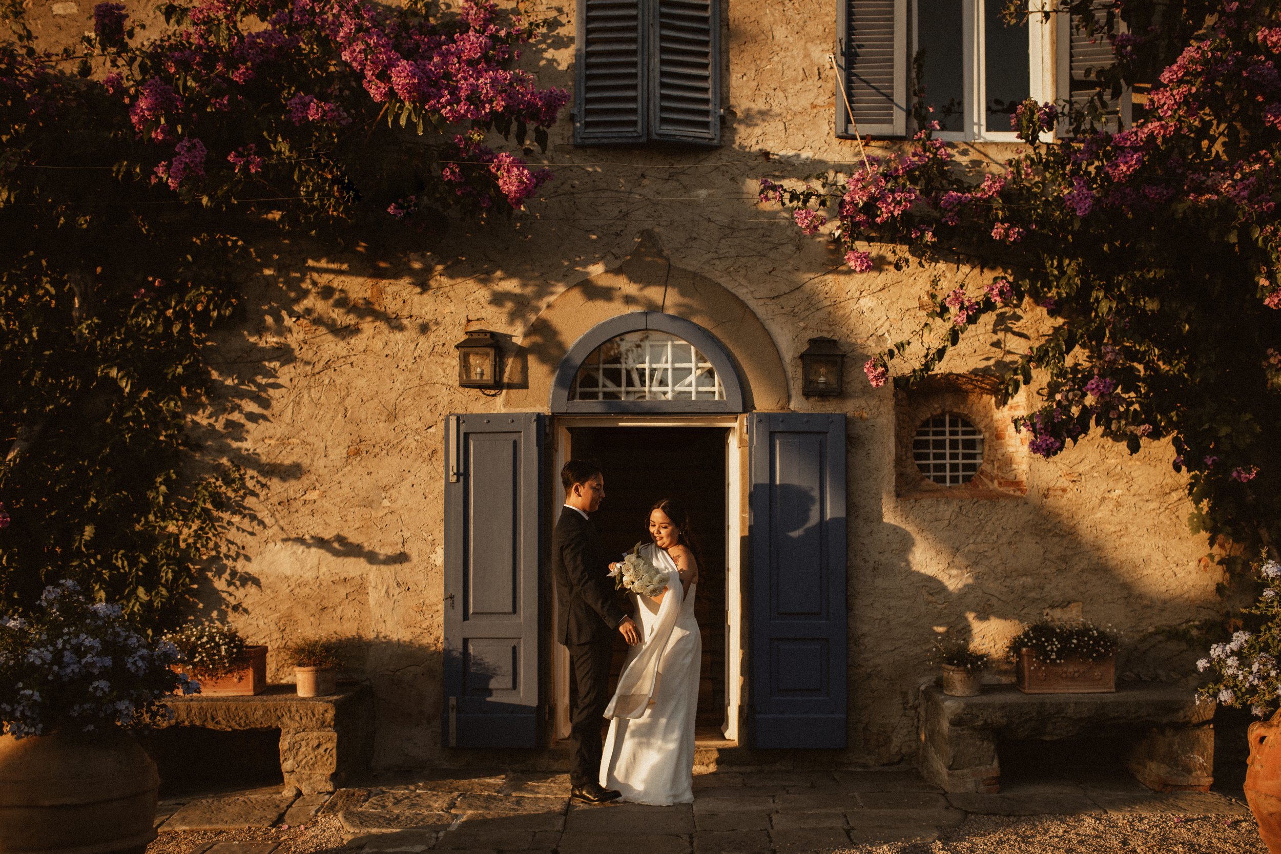A newlywed couple standing at the door of a rustic stone house with blue shutters and flowering plants, during golden hour with warm sunlight casting shadows.