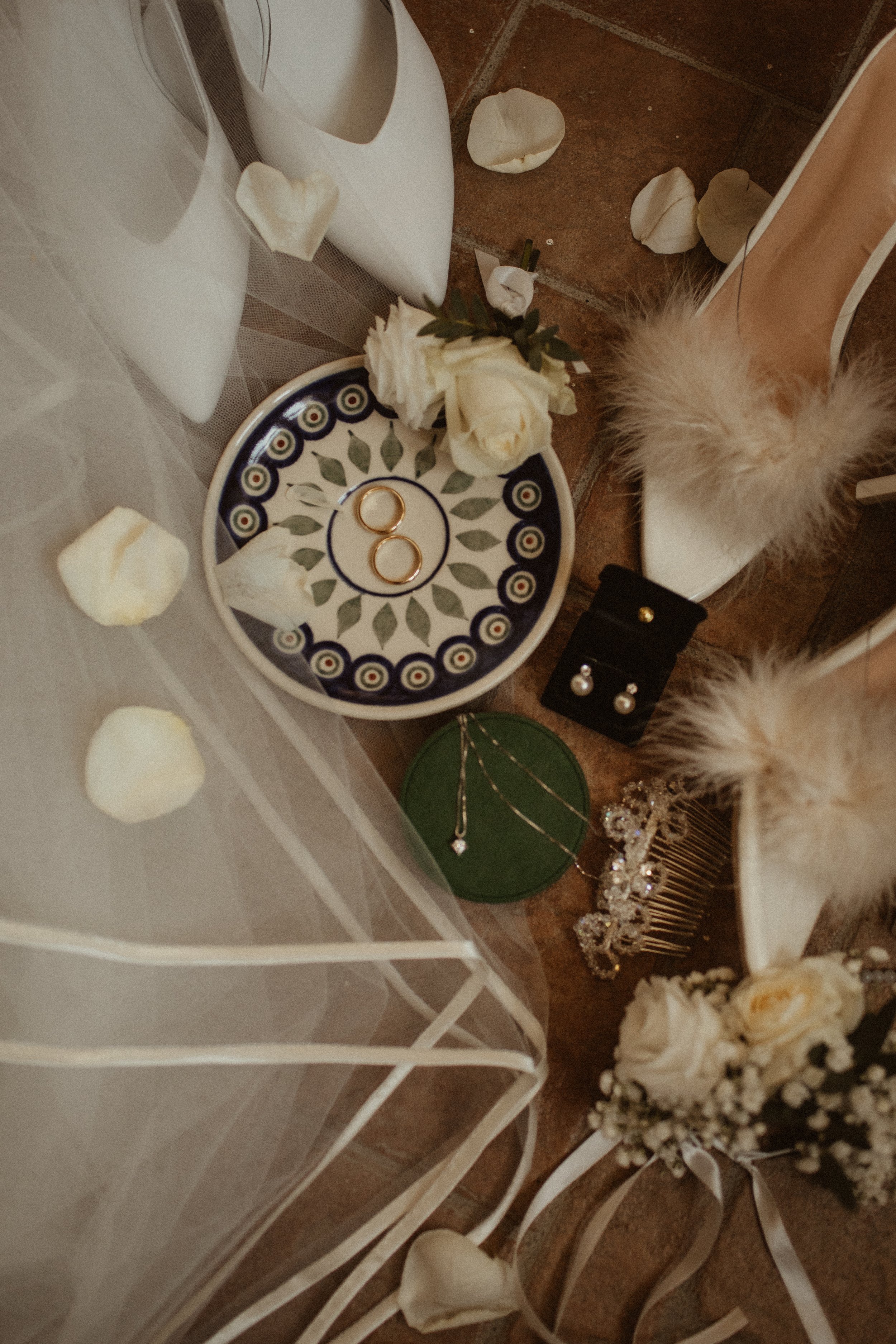 A display of wedding or engagement rings on a decorative plate, surrounded by white roses, a pair of white wedding shoes with furry accents, and jewelry including earring boxes, a hair comb, and a necklace, with scattered rose petals and flower arran