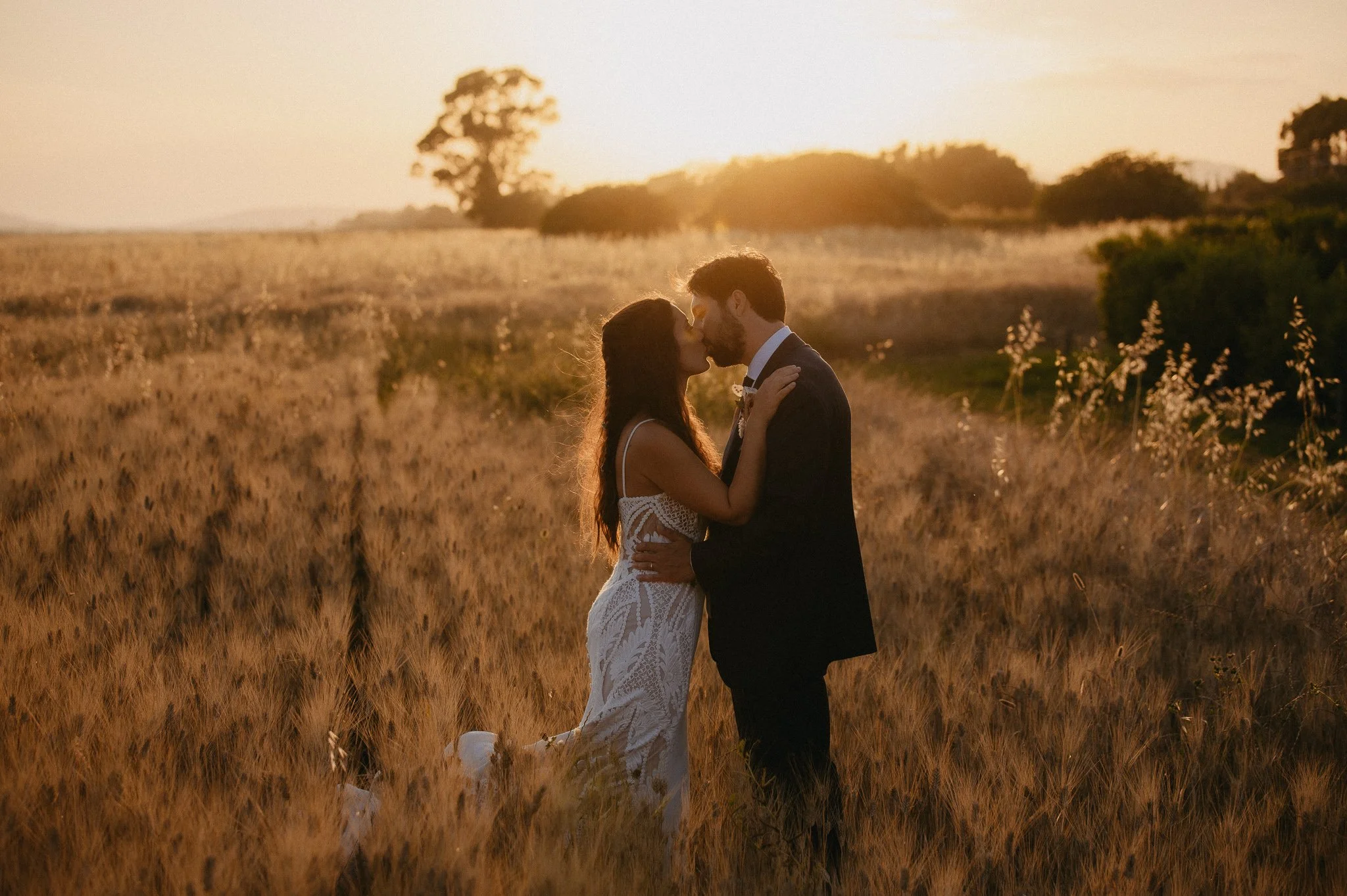 A couple kissing in a field at sunset, woman in a white lace dress, man in a dark suit.