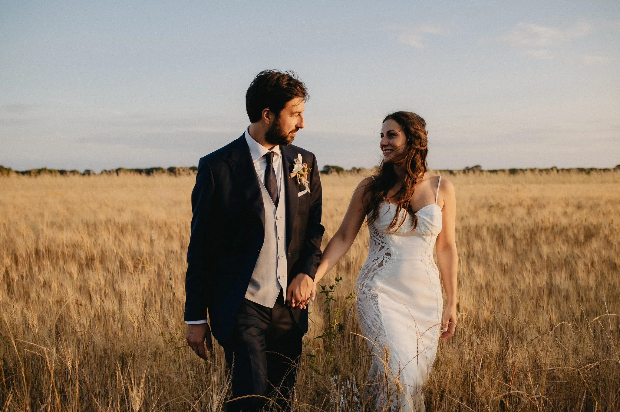 A bride and groom holding hands and walking through a golden wheat field at sunset. The groom has dark hair and beard, wearing a black suit with a white vest and a boutonniere. The bride has long brown hair, wearing a white lace wedding gown, smiling