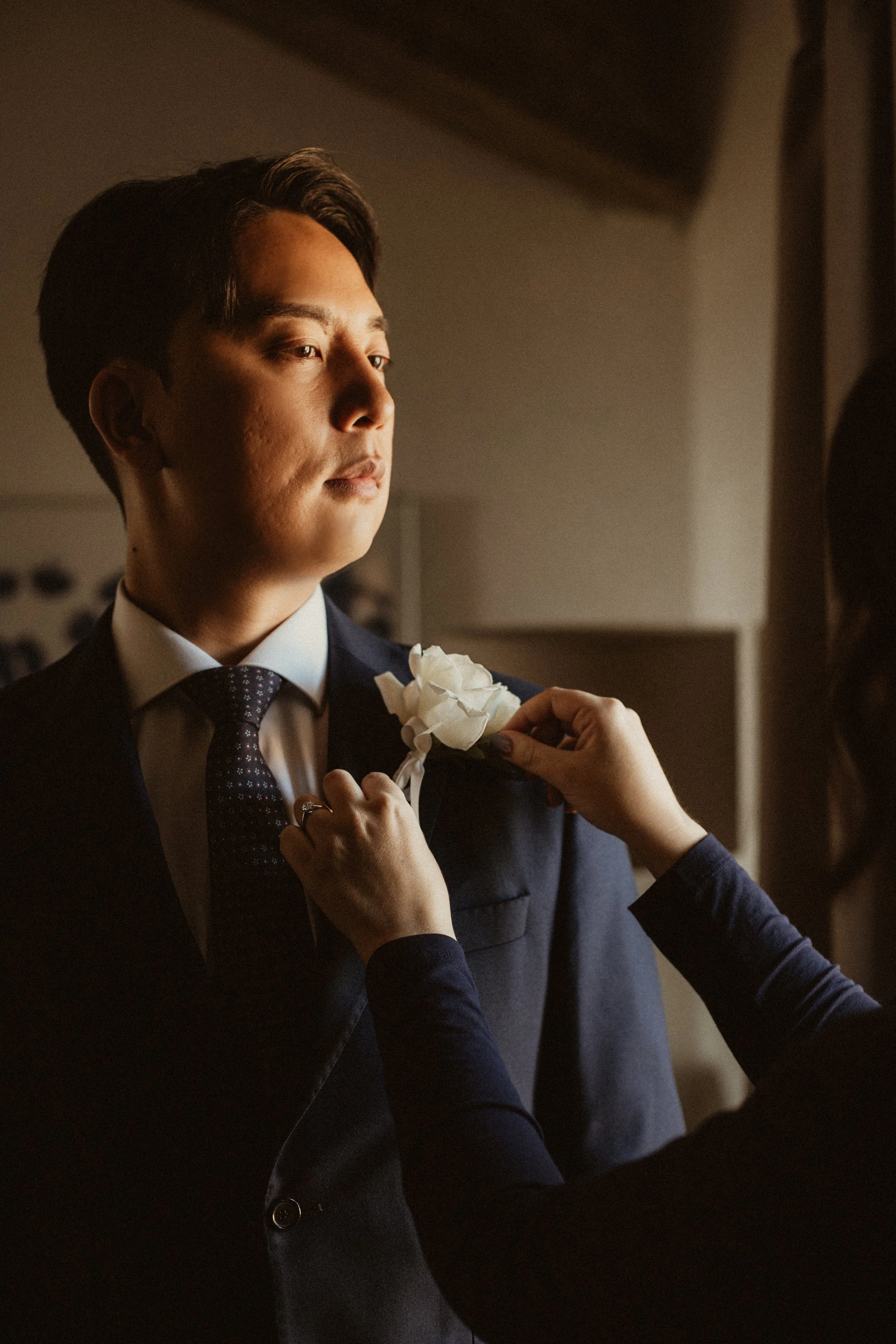 A man dressed in a suit and tie having a white flower pinned to his lapel by a woman.