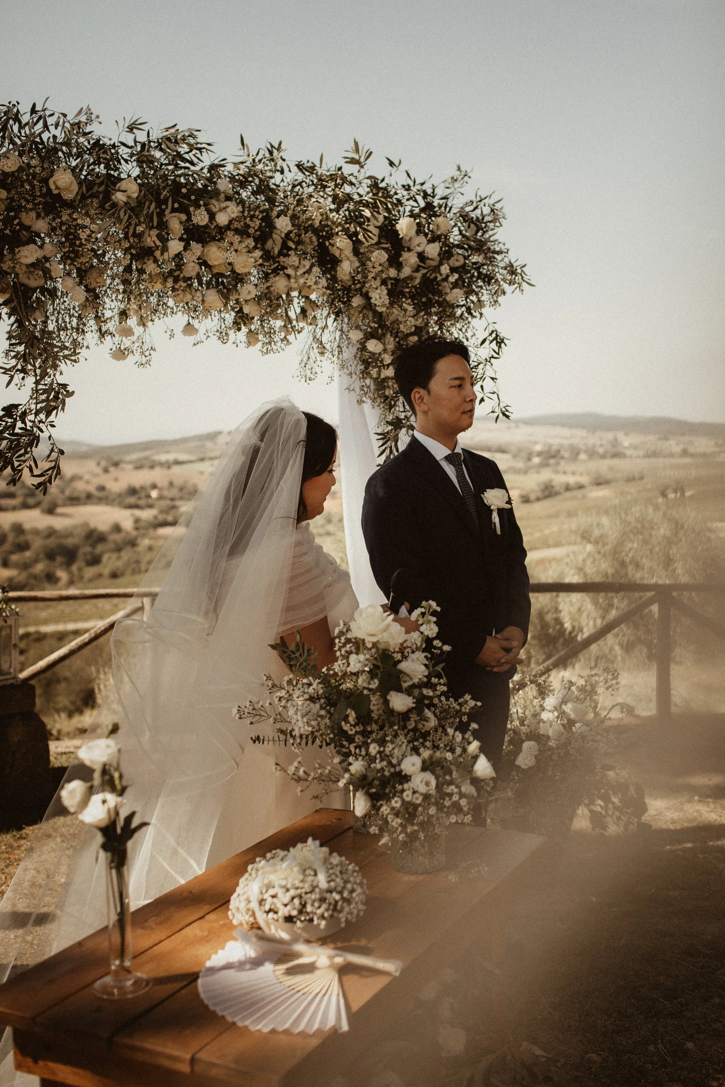 A bride and groom during their outdoor wedding ceremony beneath a floral arch, with scenic countryside in the background.
