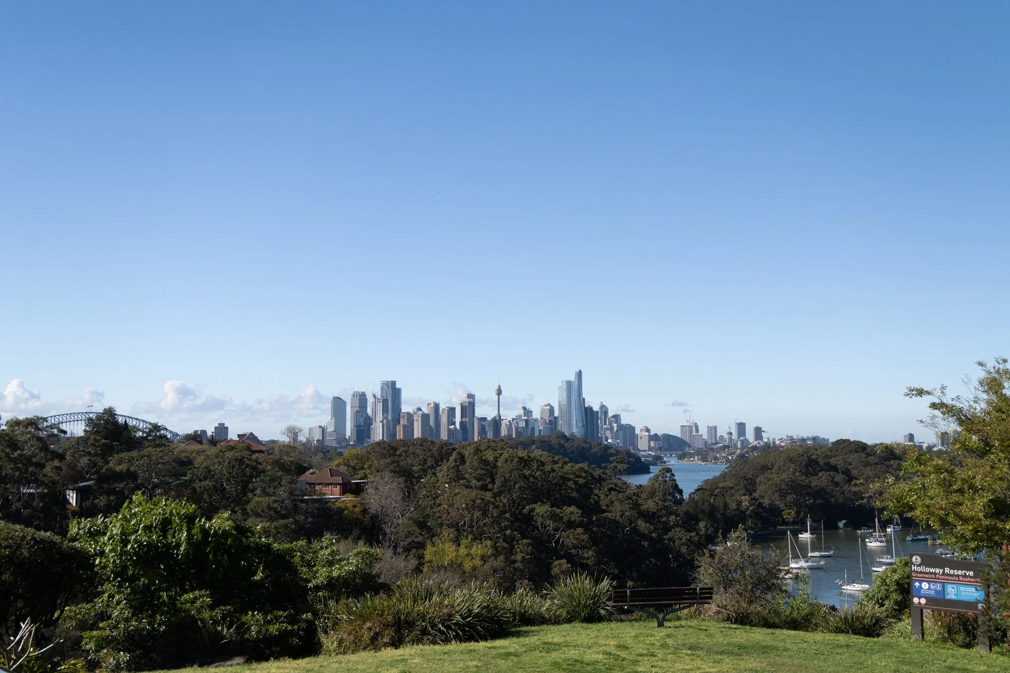 City skyline of Sydney, Australia, with tall skyscrapers near the waterfront, surrounded by trees and a park in the foreground.