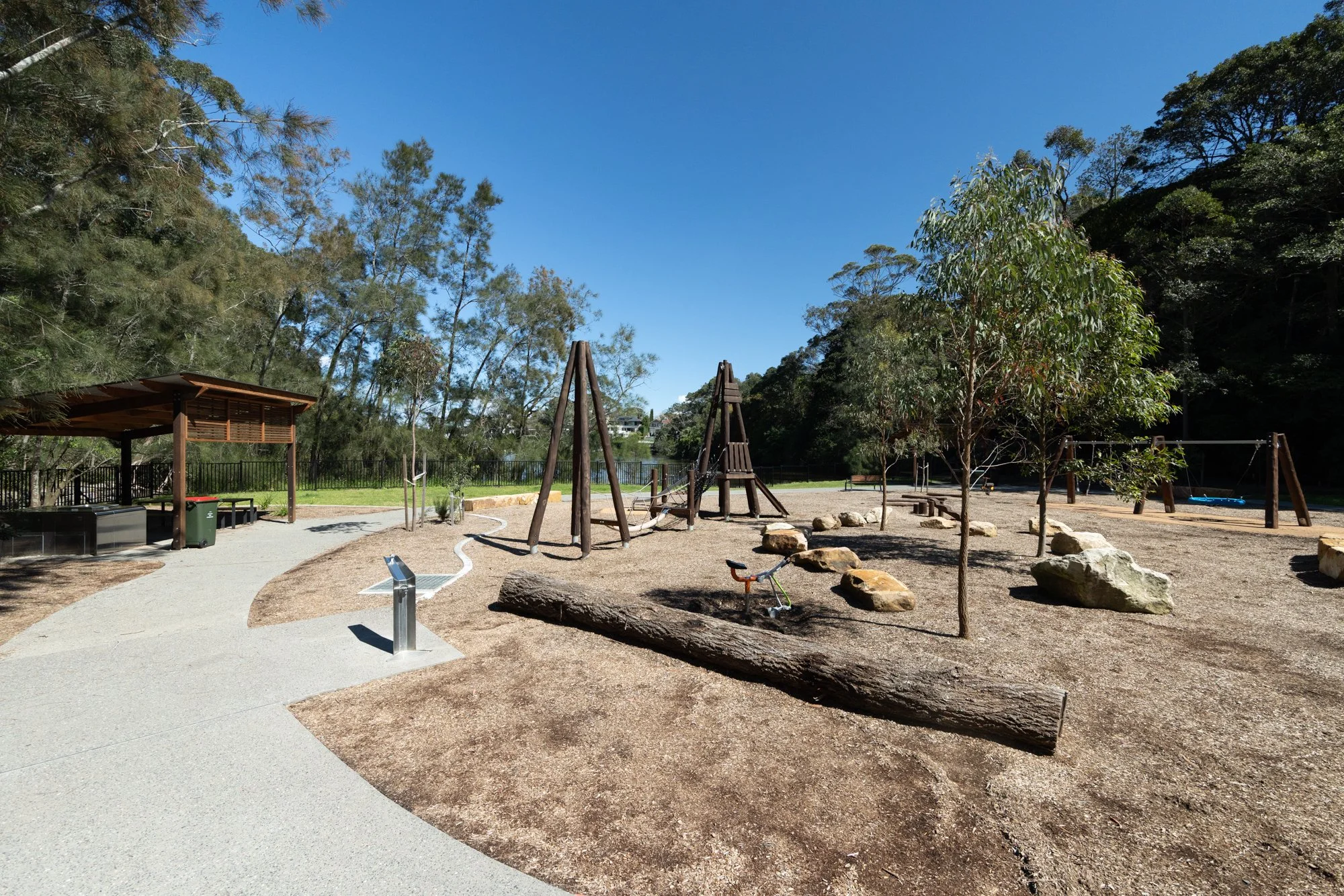 A peaceful outdoor playground with swings, trees, rocks, and a shaded picnic area under a bright blue sky.