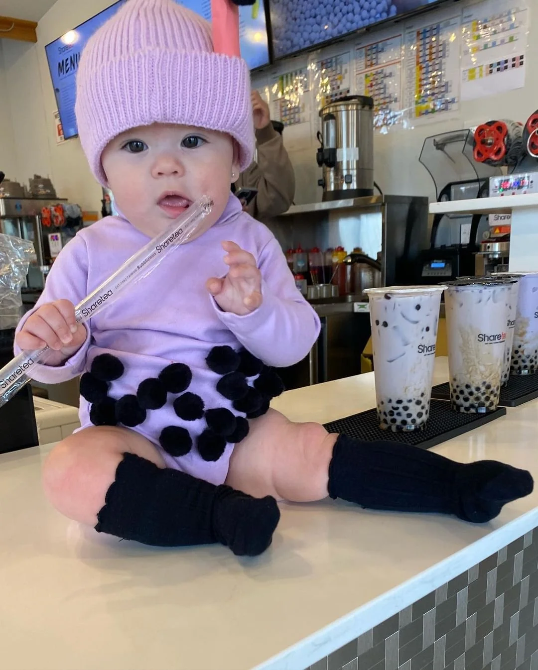 Cute baby in pink beanie and purple onesie with tongue out, holding a Sharetea straw at a boba tea shop counter with drinks.