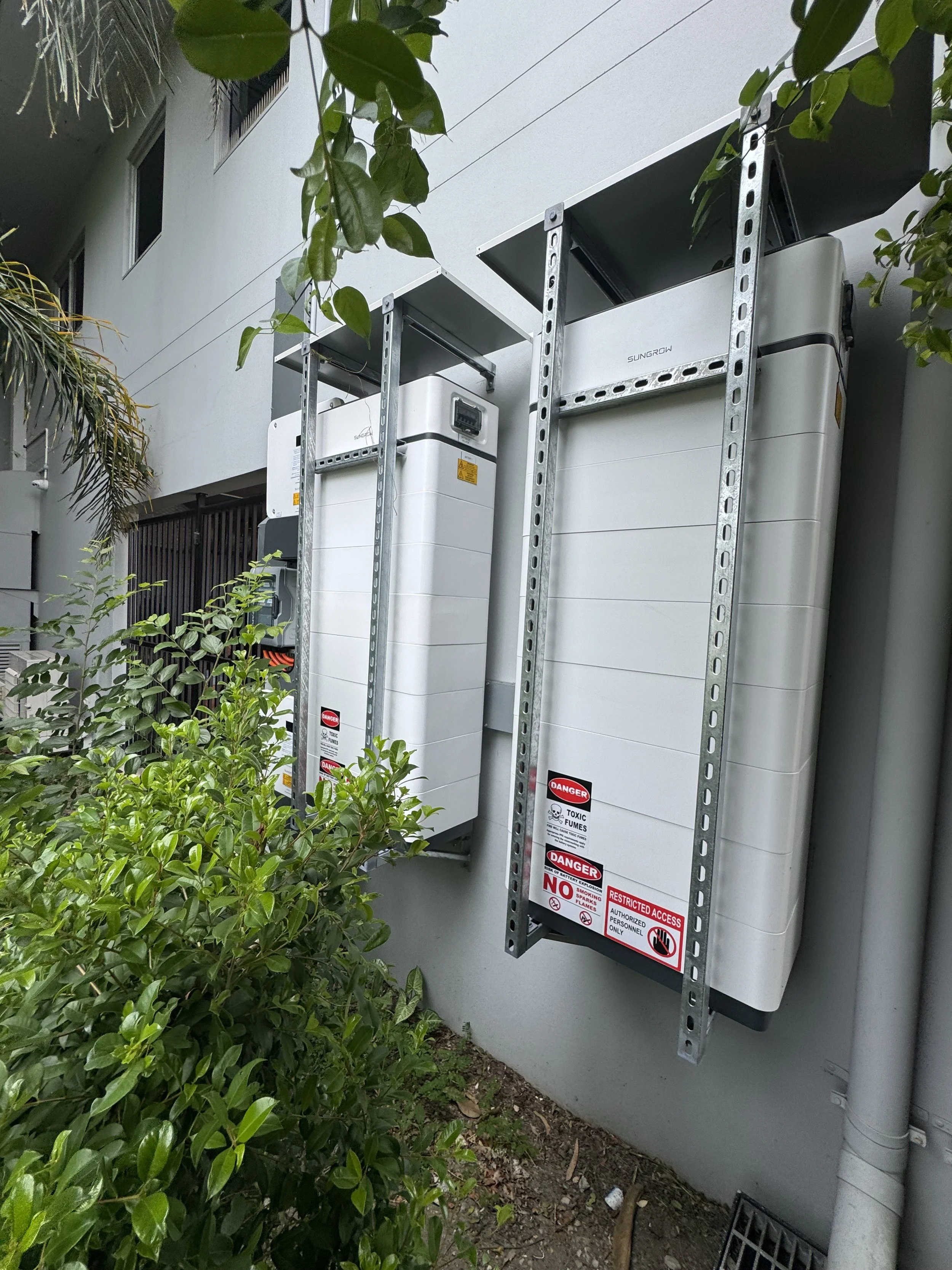 Two utility meters, possibly for electricity, installed on an exterior wall with metal supports. Green bushes in the foreground and part of a building with windows above.