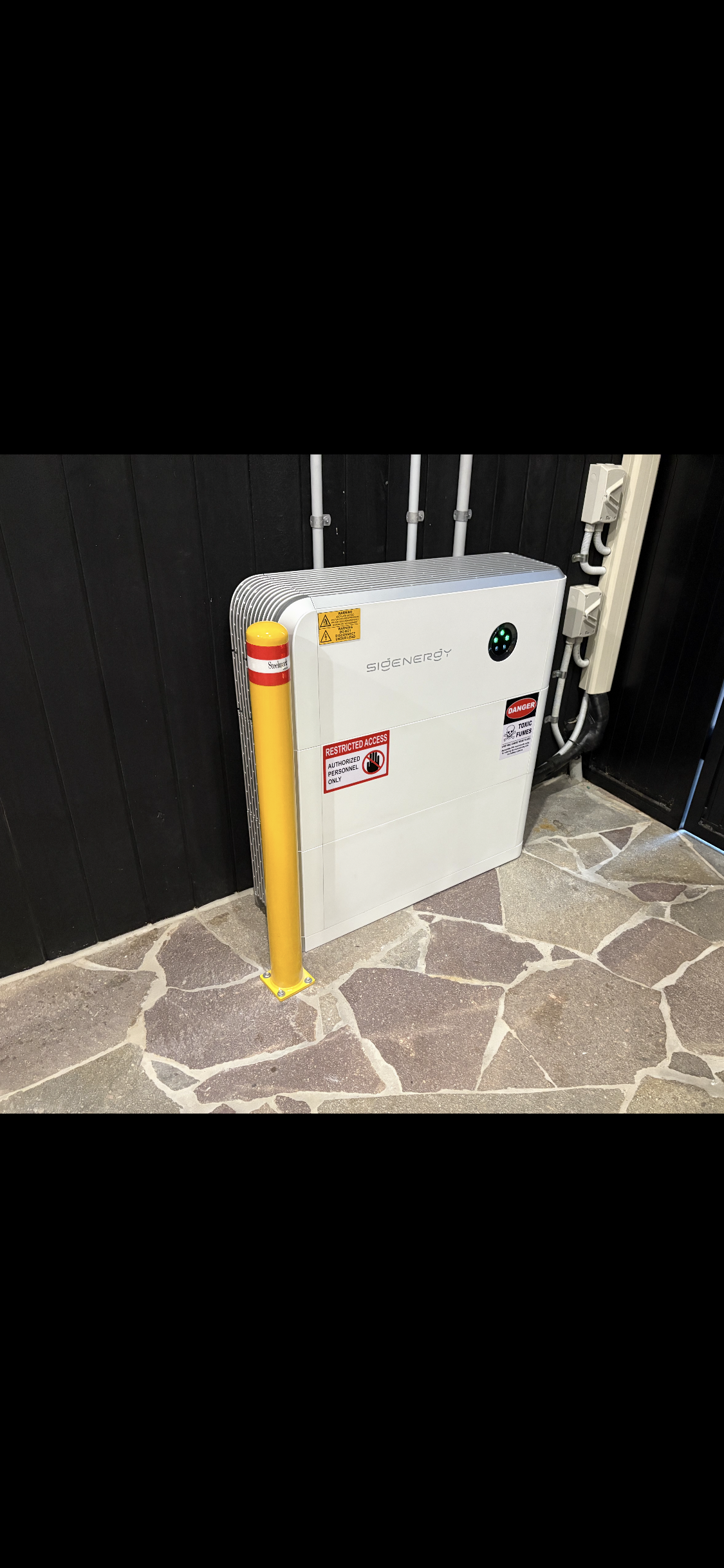 A white Sigenergy electric vehicle charging station with warning and restricted access signs, positioned on a stone-paved floor next to a black wall with electrical conduits.