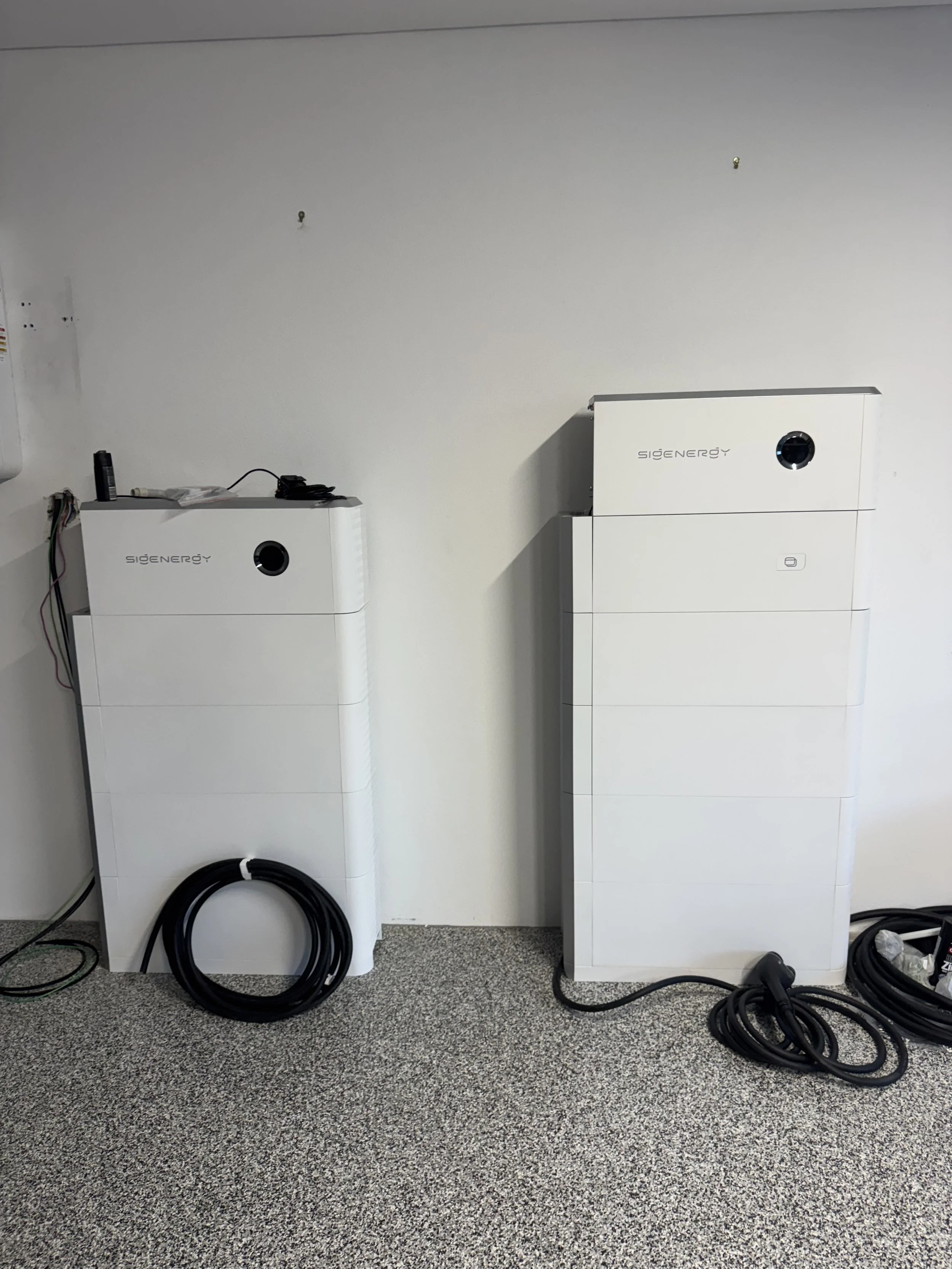 Two white energy storage systems labeled 'SigEnergy' are standing on a carpeted floor against a plain wall. The left unit has some wires on top and a coil of pipe at the bottom, while the right unit has a small display and a round opening.