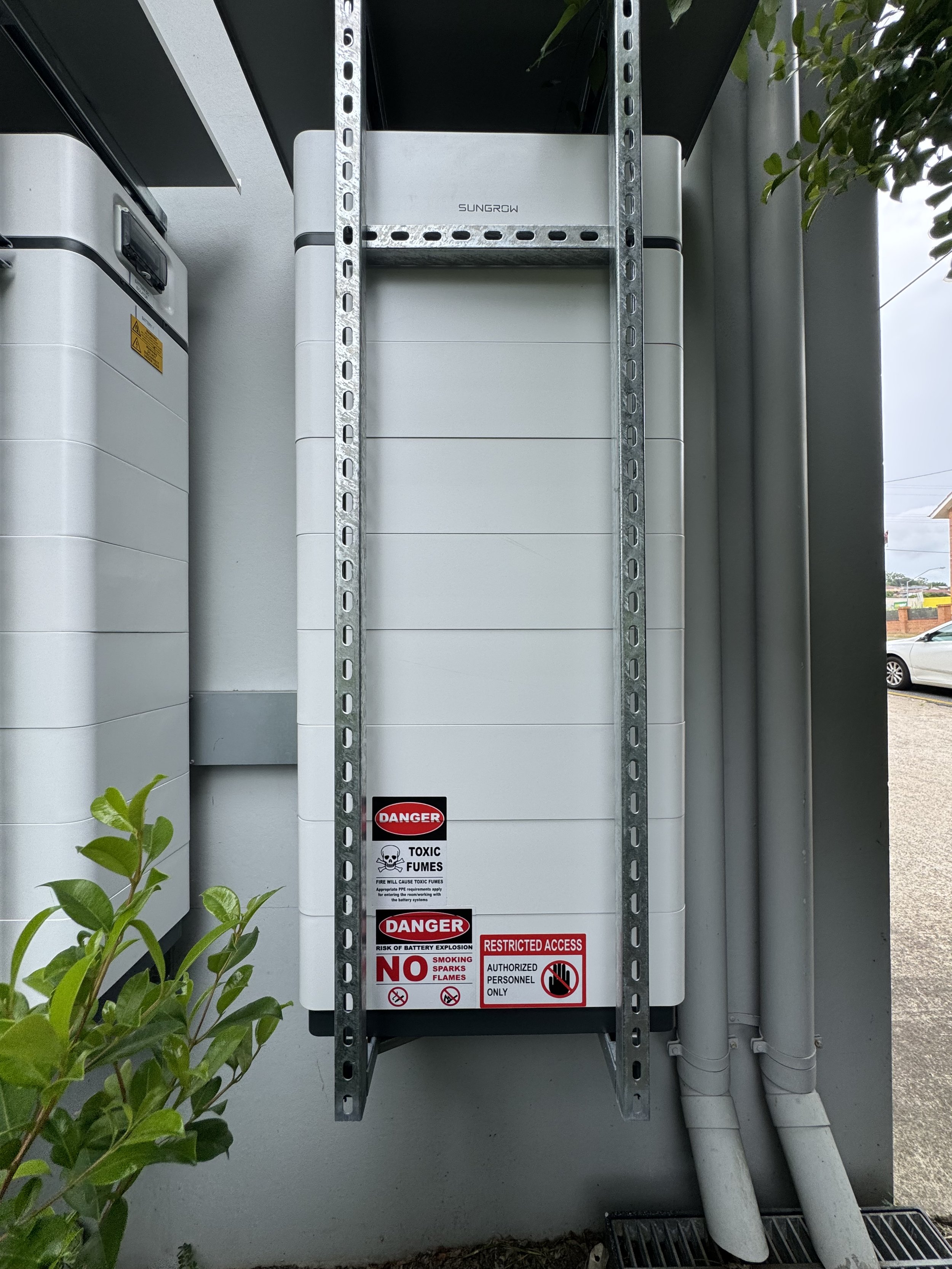 Electrical equipment enclosure with warning labels, surrounded by pipes and a plant, outside.