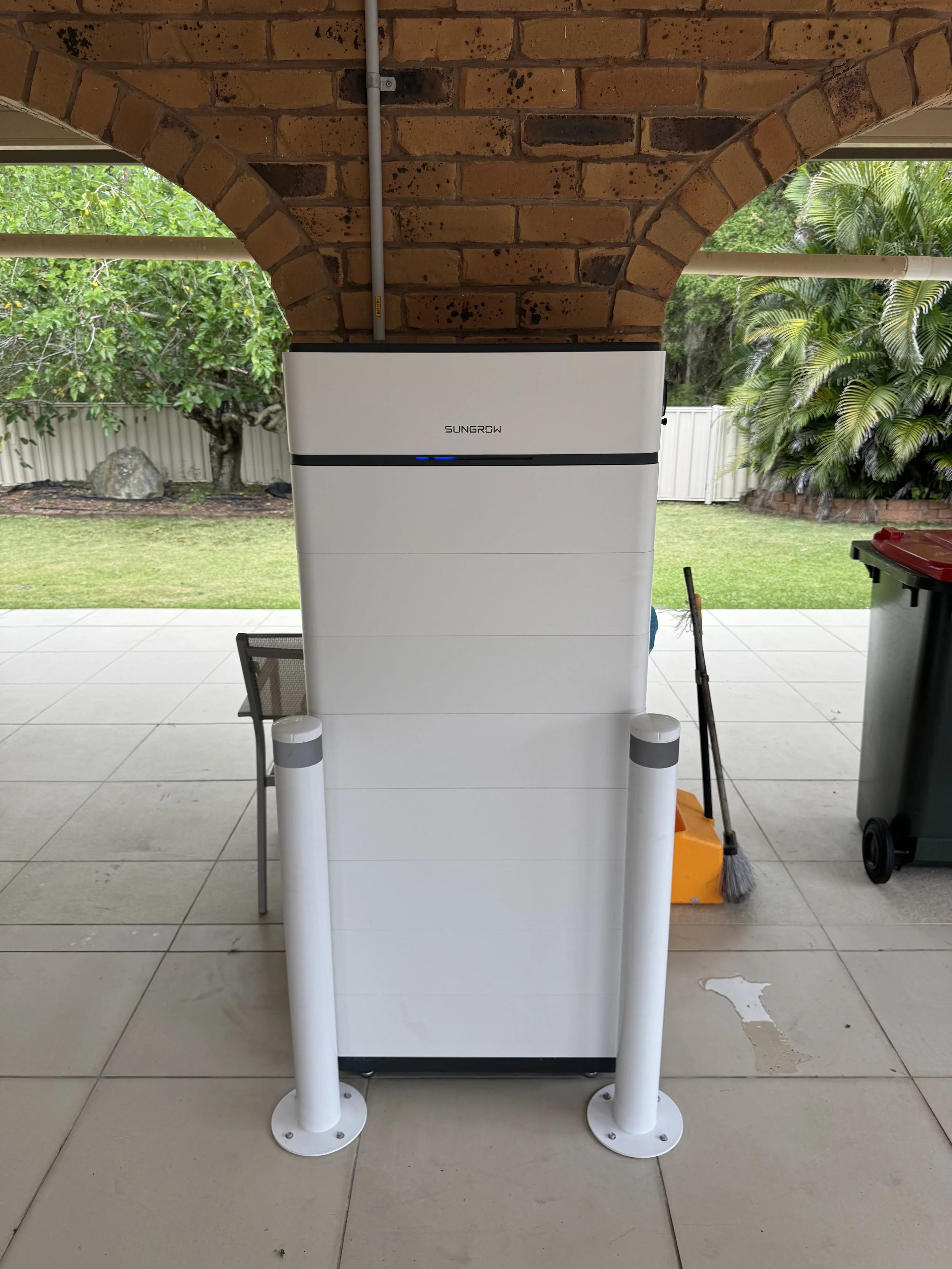 An outdoor view of a white SunGrow solar inverter installed under a brick archway, with a garden and trees in the background.