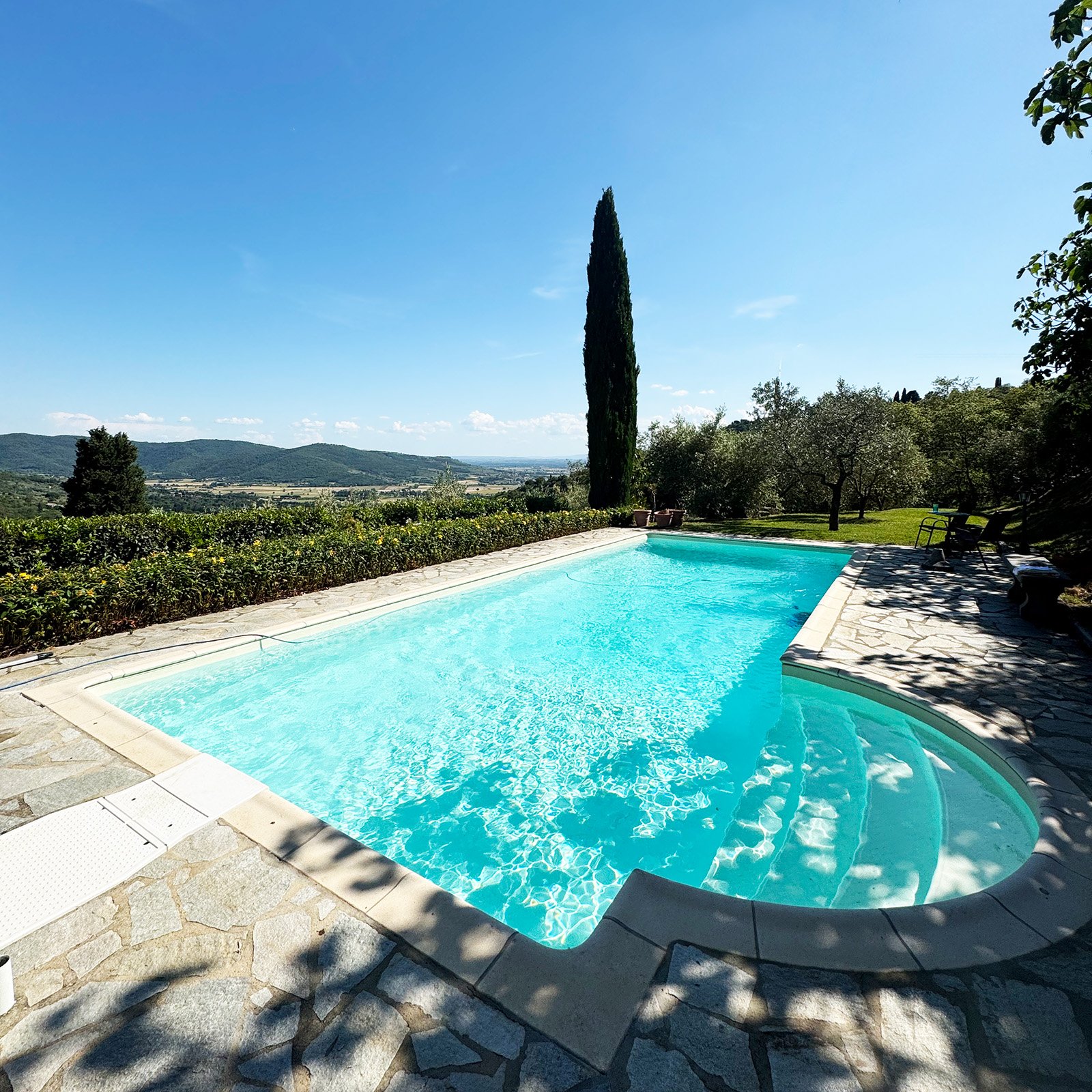 A swimming pool surrounded by stone paving with a scenic countryside view in the background, features a tall cypress tree, outdoor seating, and shaded areas.