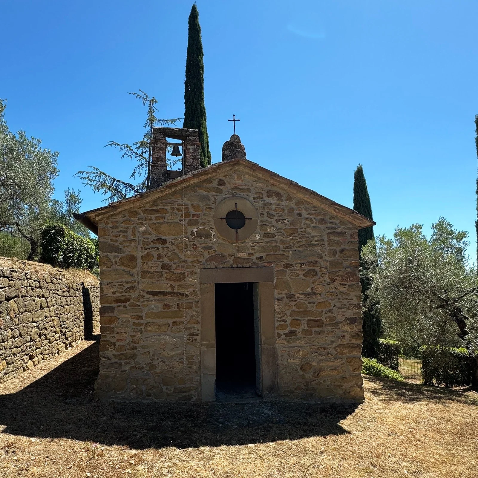 Small stone church with open door, bell tower, cross on top, surrounded by cypress and olive trees, under clear blue sky.