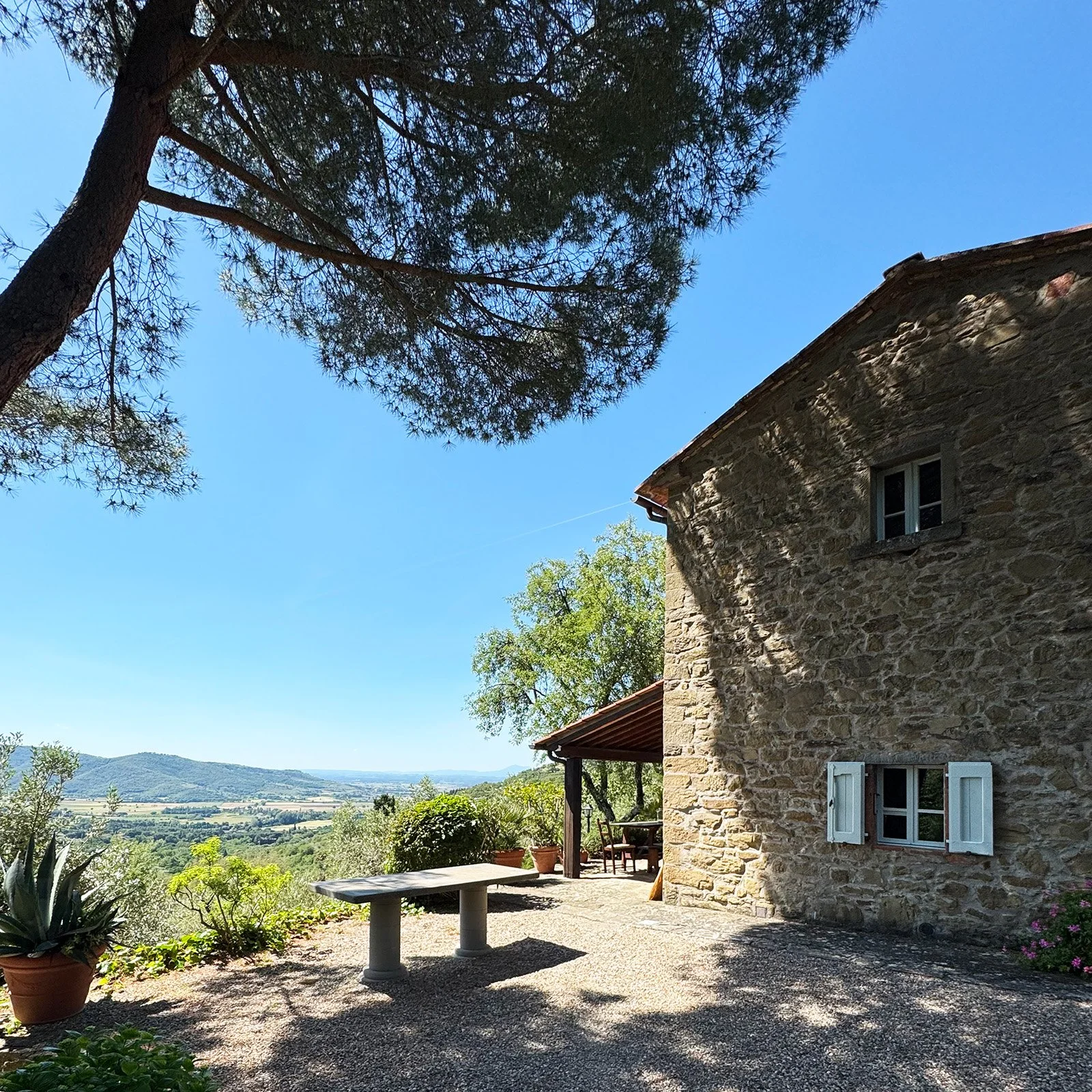 Stone house with white window shutters on a sunny day, overlooking a landscape with trees and mountains in the distance.