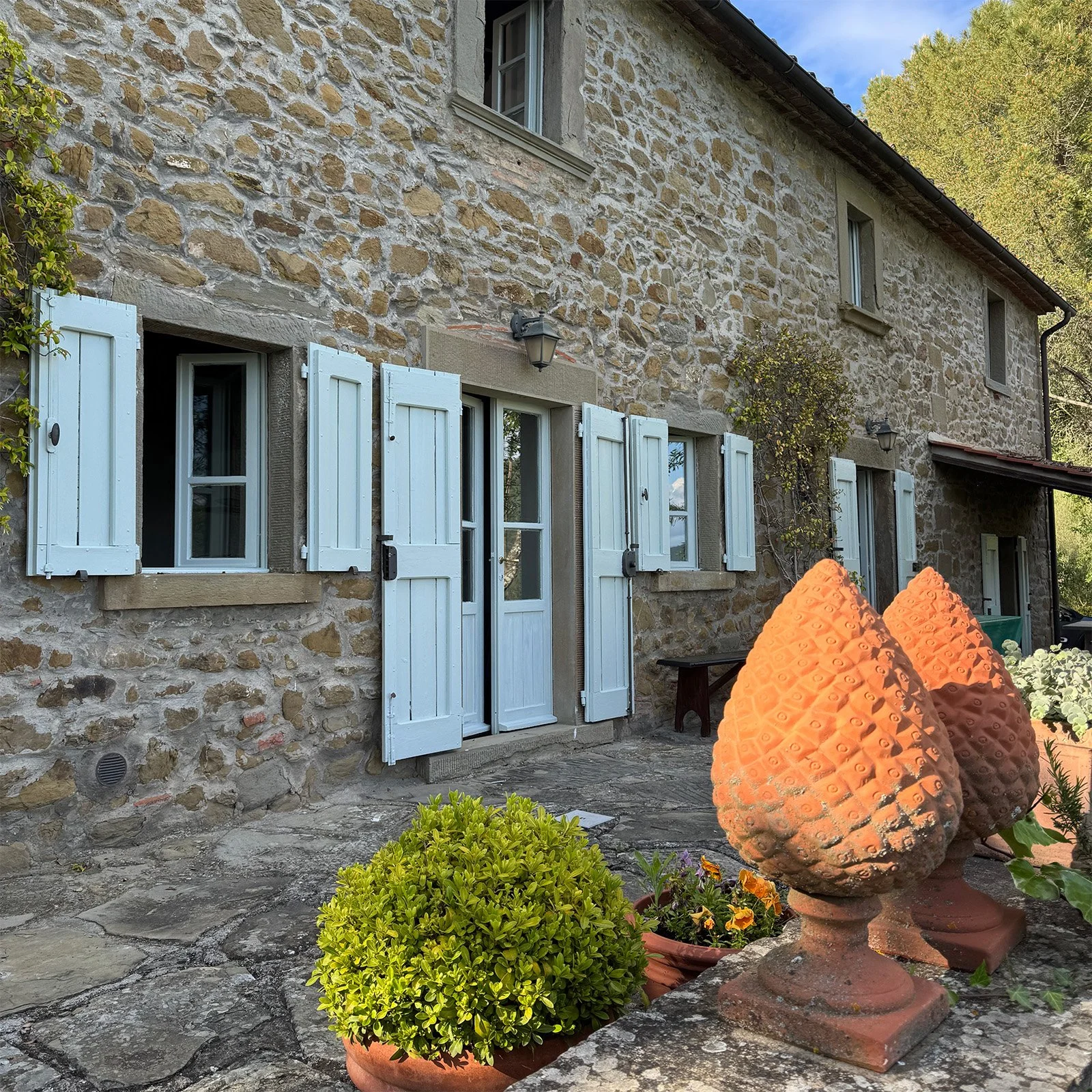 Stone house with white window shutters and potted plants on a stone patio.