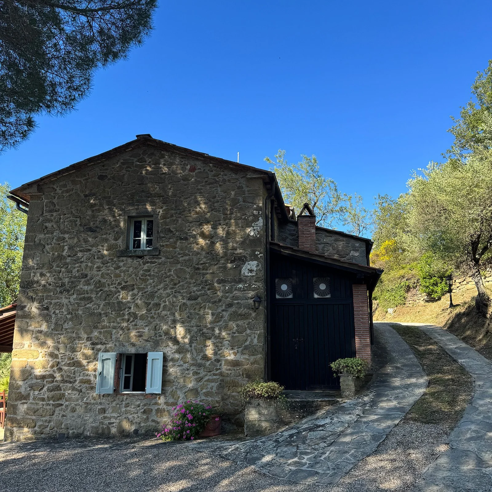 A rustic stone house with white window shutters, a black garage door, and a stone driveway, surrounded by greenery under a clear blue sky.