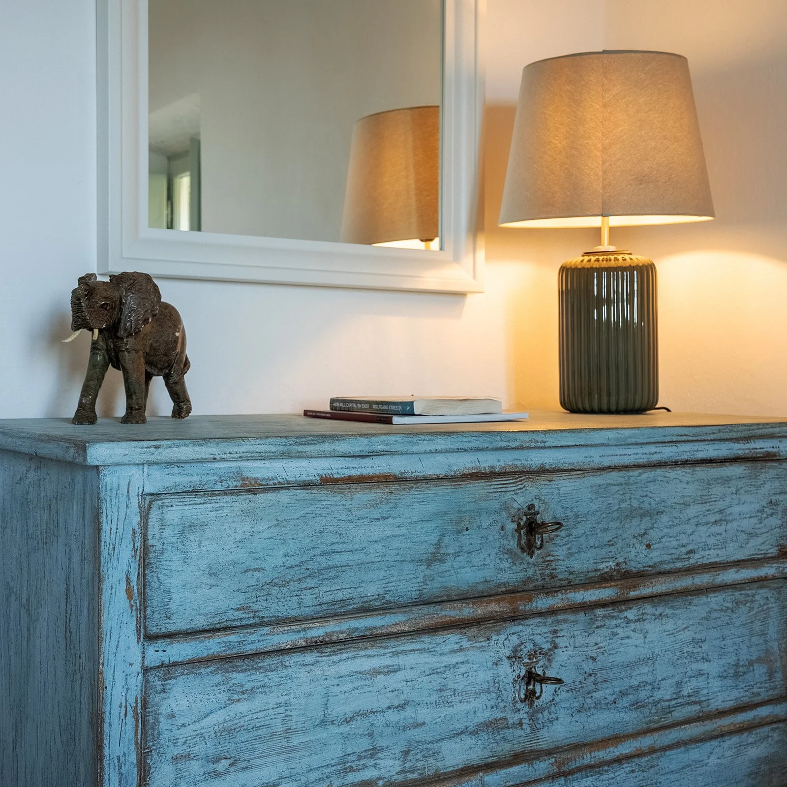 A vintage blue distressed wooden dresser with a small bronze elephant figurine, a few books, and a lamp with a beige shade. A mirror hangs above the dresser, reflecting part of the room.
