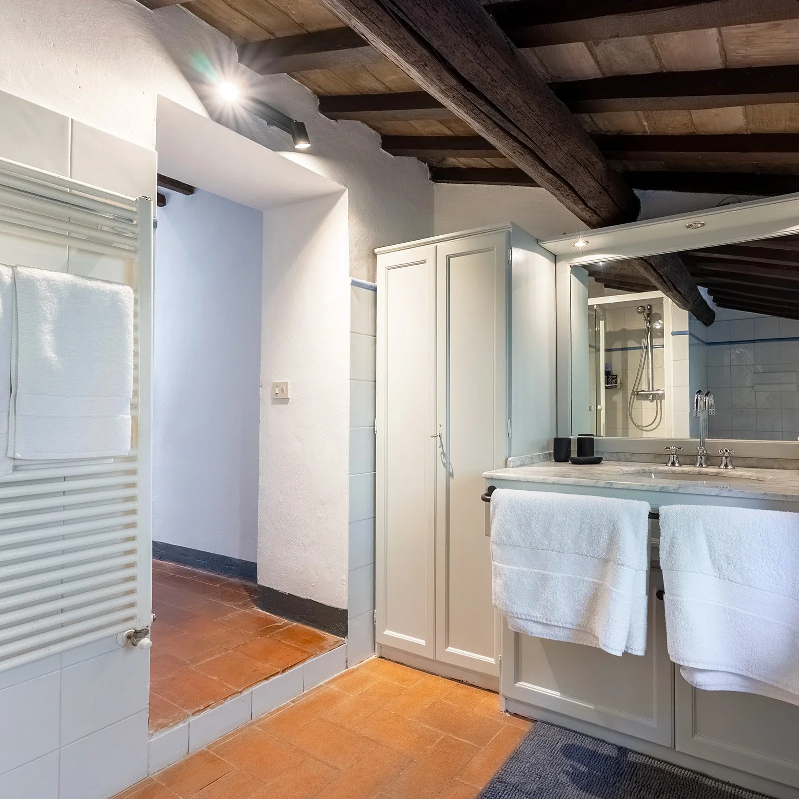 A bathroom with a wooden ceiling, white walls, and a terracotta tiled floor. There is a large mirror above a stone countertop with a sink, two white towels hanging from the front, and black containers on the counter. To the left, a white radiator wit