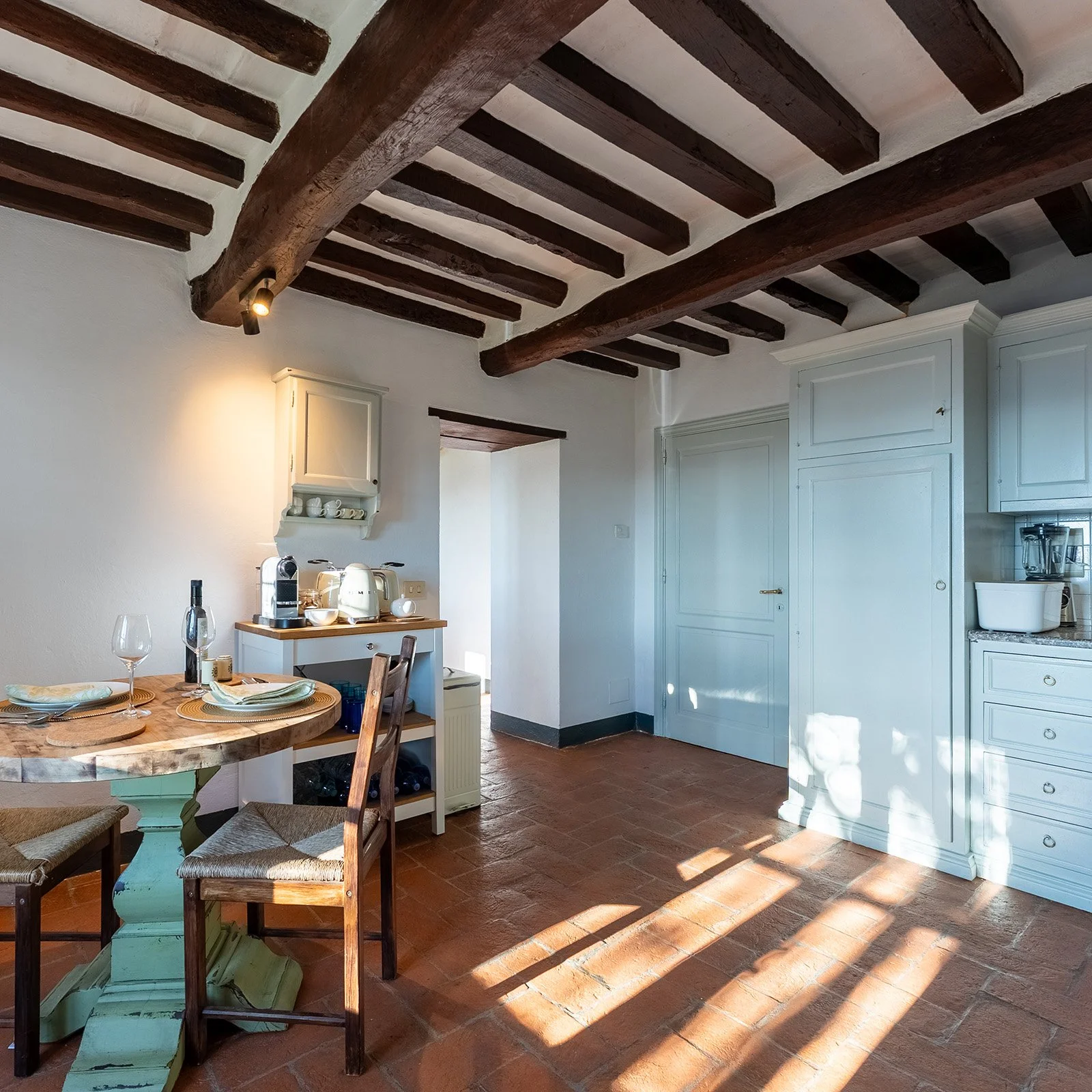 A cozy kitchen with white cabinetry, wooden beams on the ceiling, a round dining table with chairs, and sunlight streaming through windows casting shadows on the terracotta tile floor.