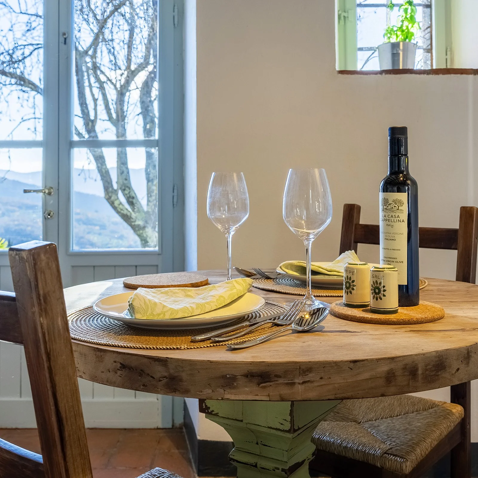 A round wooden dining table set with two empty wine glasses, two plates with yellow napkins, a cork coaster, and silverware. There are also a bottle of olive oil, two small salt and pepper shakers, and a small potted plant on a circular cork mat. In 
