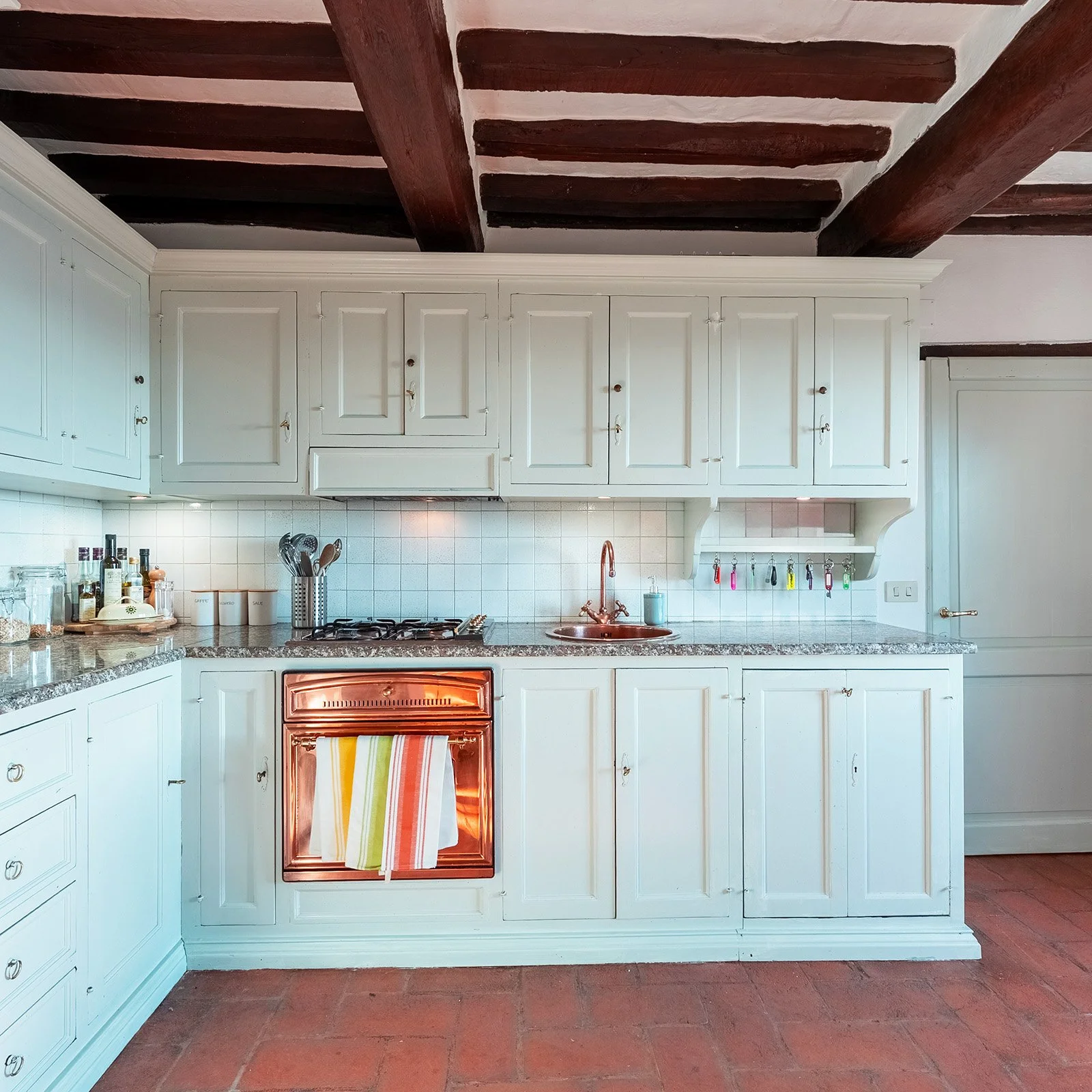 Kitchen with white cabinets, granite countertops, a copper sink, and a copper oven with colorful towels hanging. Wooden ceiling beams and a tiled backsplash. Various kitchen items and utensils are on the counters.