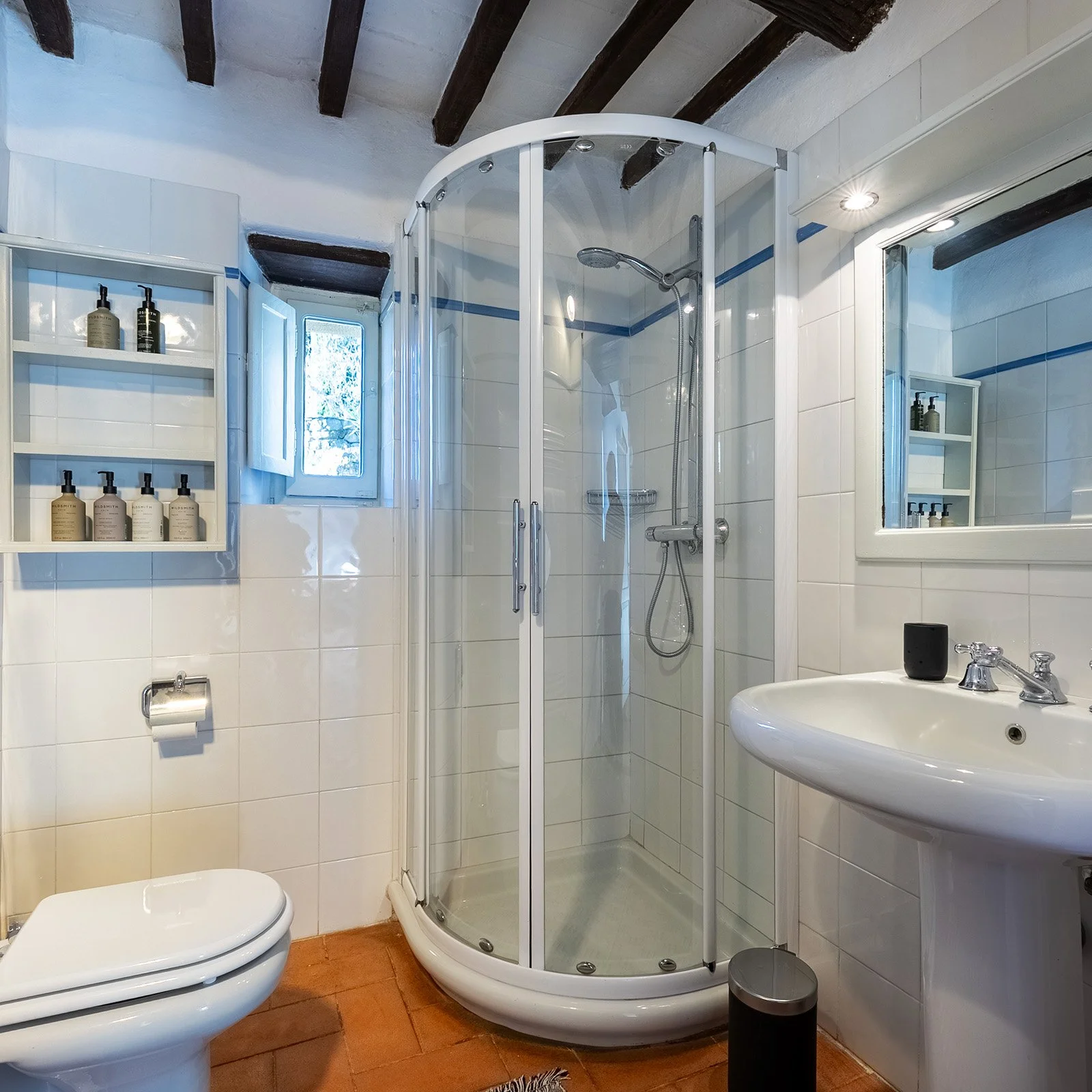 A small bathroom with a corner shower stall with glass doors, a white toilet, a white sink, a mirror, a window, and built-in shelves with bottles, wooden ceiling beams, and a terracotta tile floor.