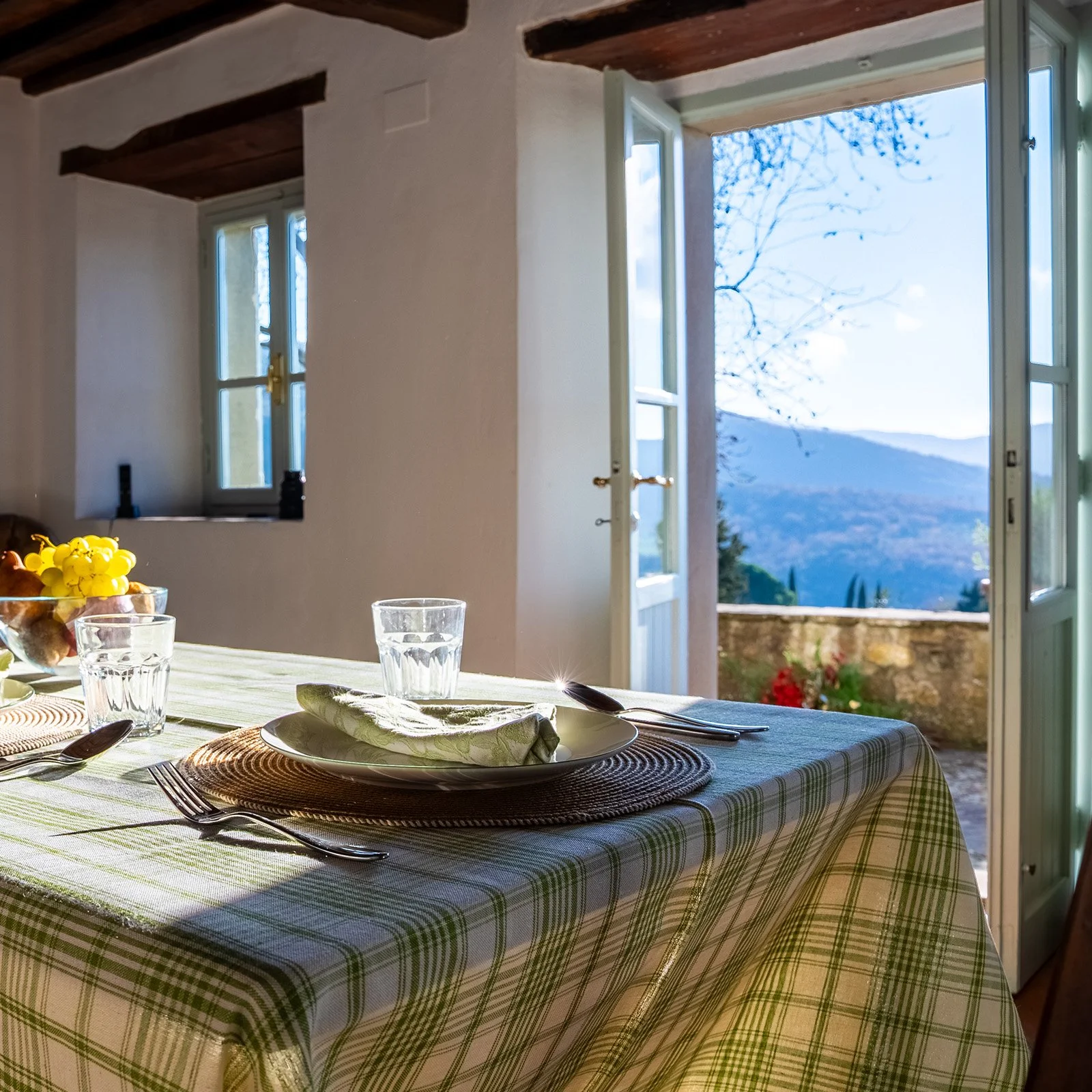 A dining room with a table set for a meal, featuring plates, glasses, silverware, and a cloth napkin. The tablecloth is green and white checkered. Bright sunlight enters through open French doors and a window, revealing a scenic view of mountains and